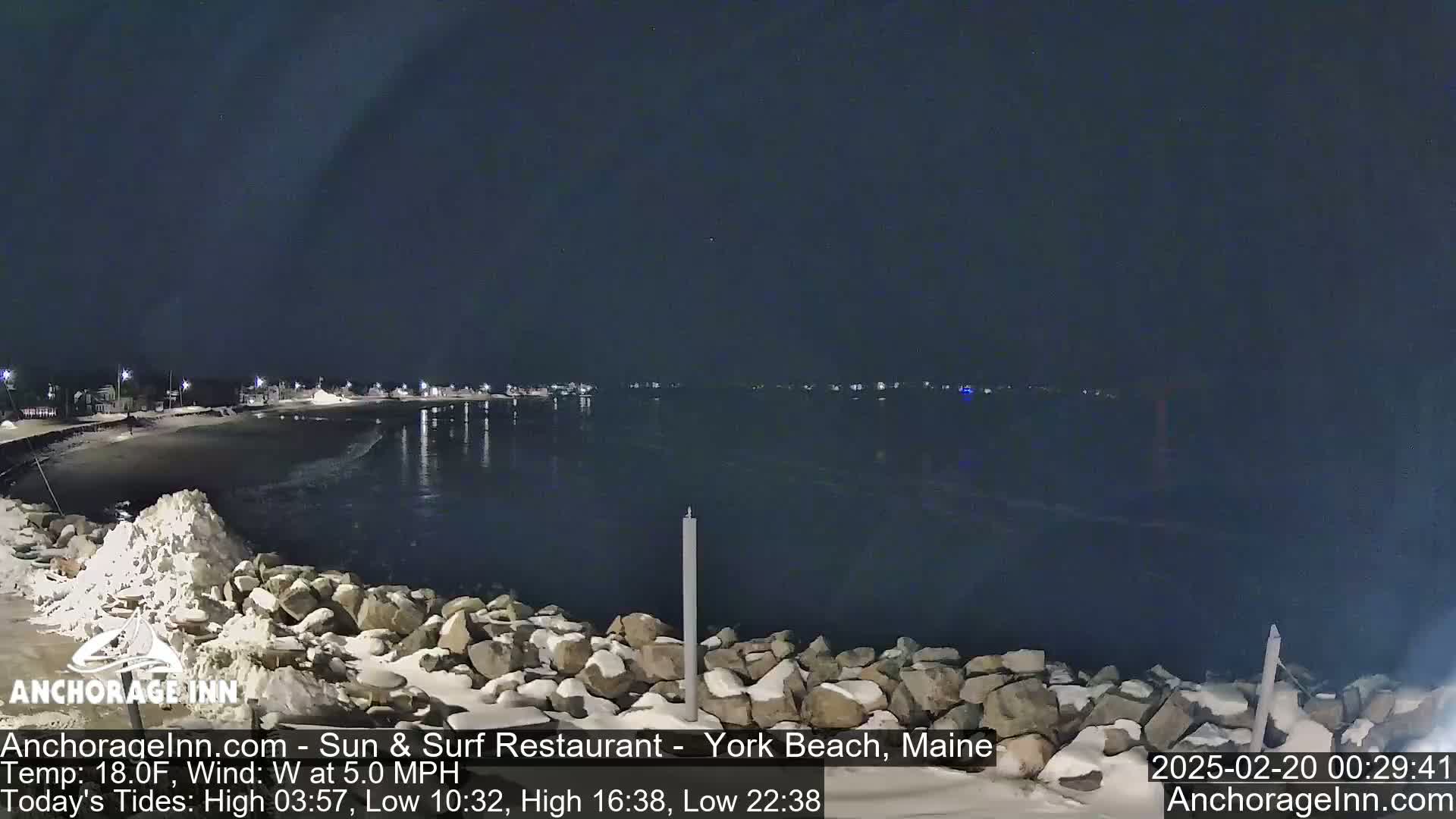 A nighttime view of a snow-dusted rocky shoreline and calm dark water, with a village of lights visible in the distance under a clear, dark sky.