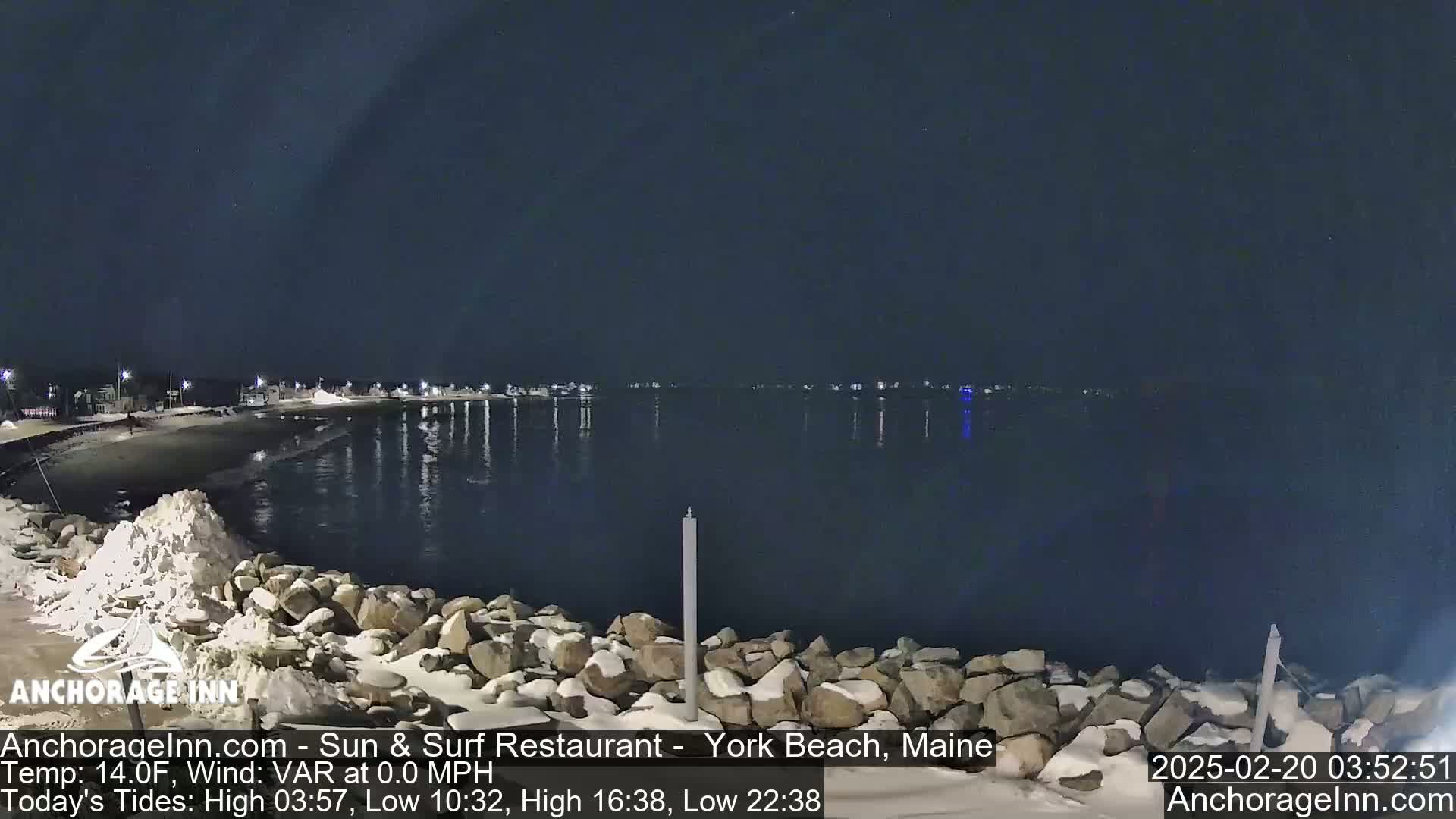 A nighttime view of a snow-dusted rocky shoreline and calm, dark water, with lights from buildings visible across the water under a clear, dark sky.