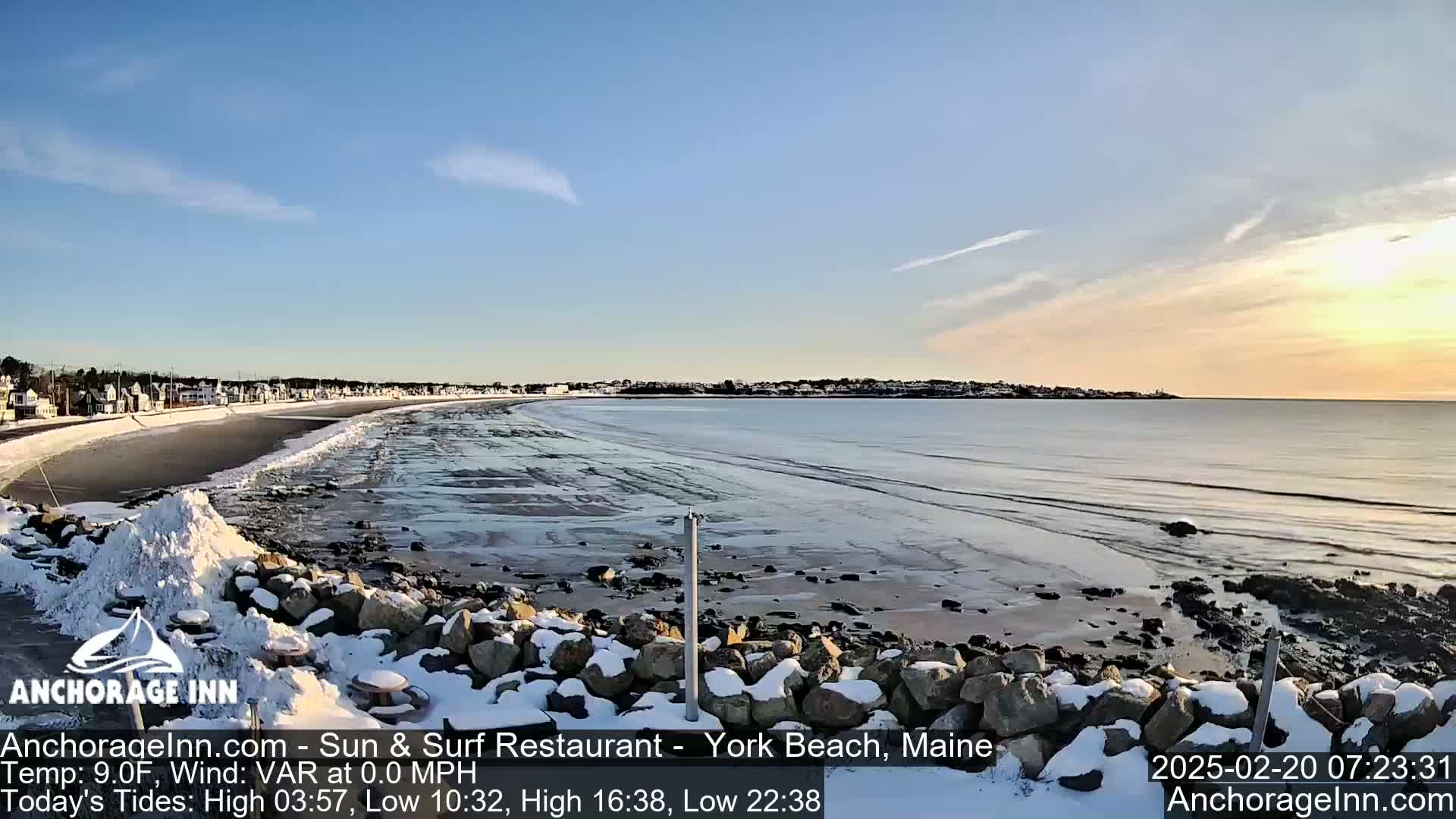 A snow-covered rocky shoreline overlooks a calm, partially frozen ocean with a town visible in the distance under a mostly clear sky.