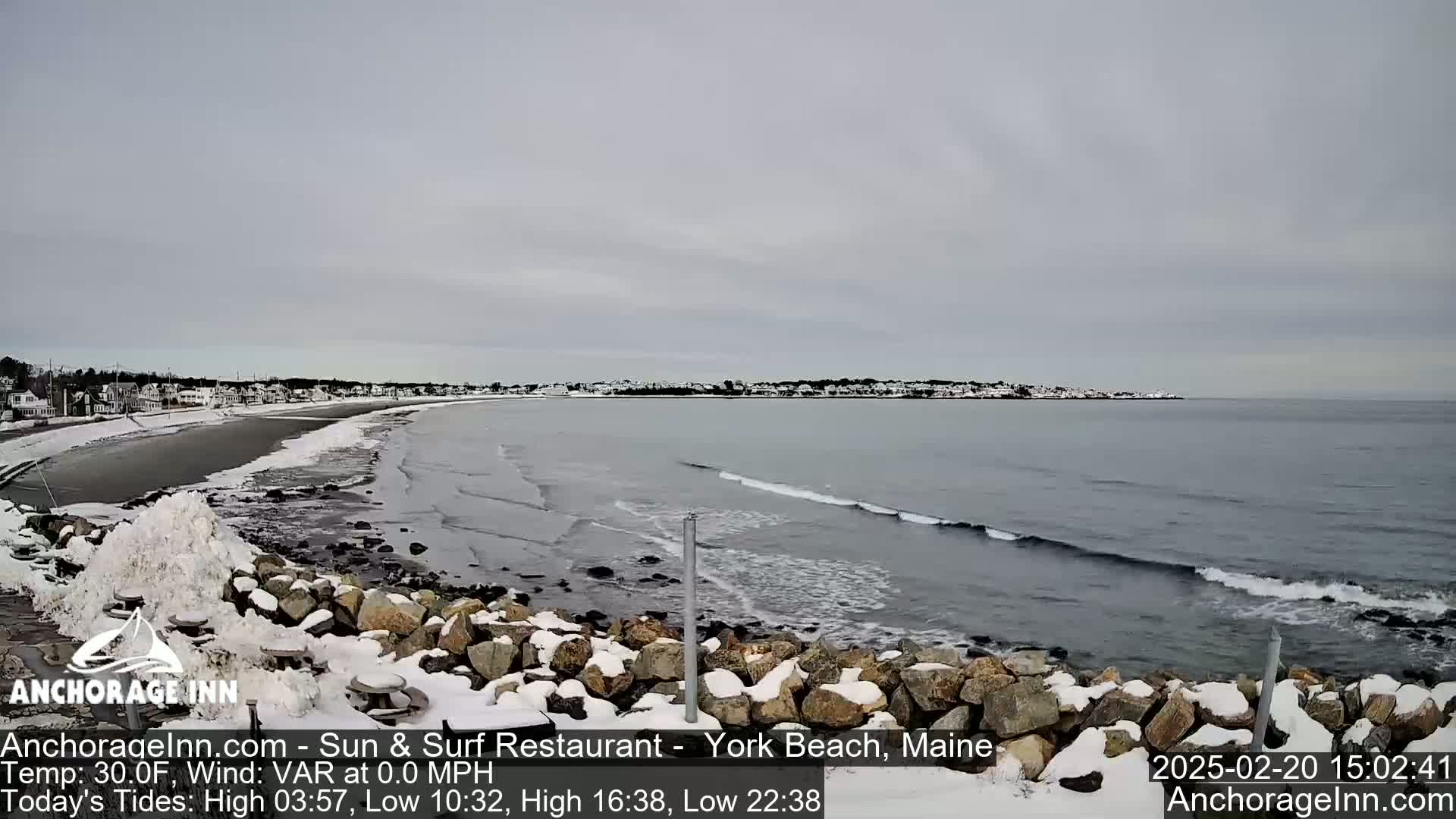 A snow-covered rocky shoreline overlooks a calm, grey ocean with small waves under an overcast sky.