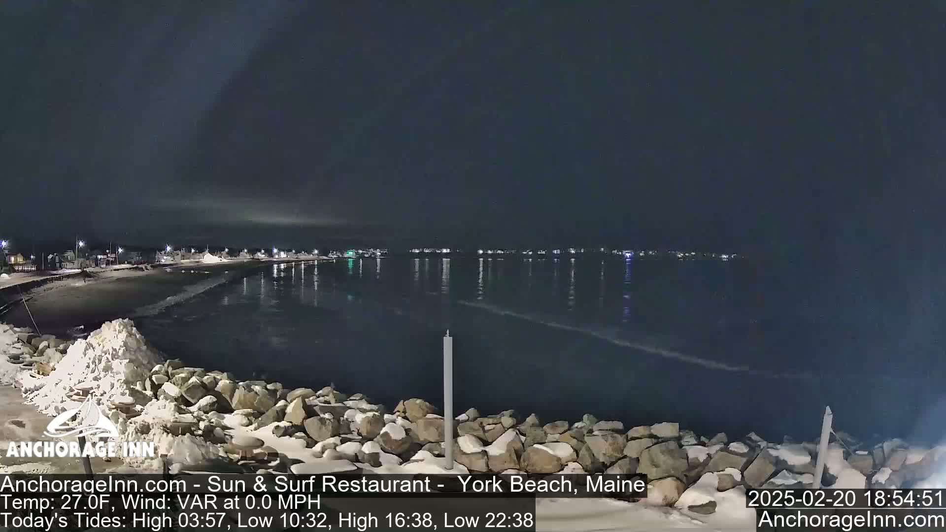 A snow-covered rocky shoreline at night overlooks a calm, dark ocean reflecting the lights of a distant town under a cloudy sky.