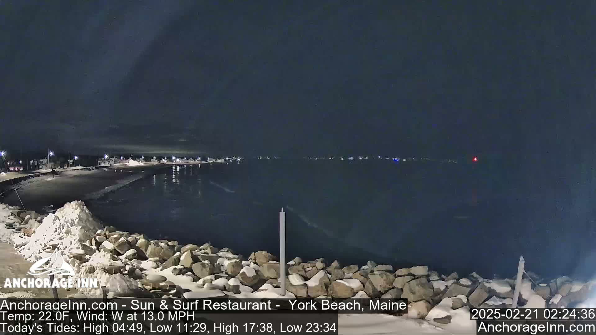 A nighttime view of a snow-covered rocky shoreline and calm, dark water, with a town faintly lit in the distance under a dark, clear sky.