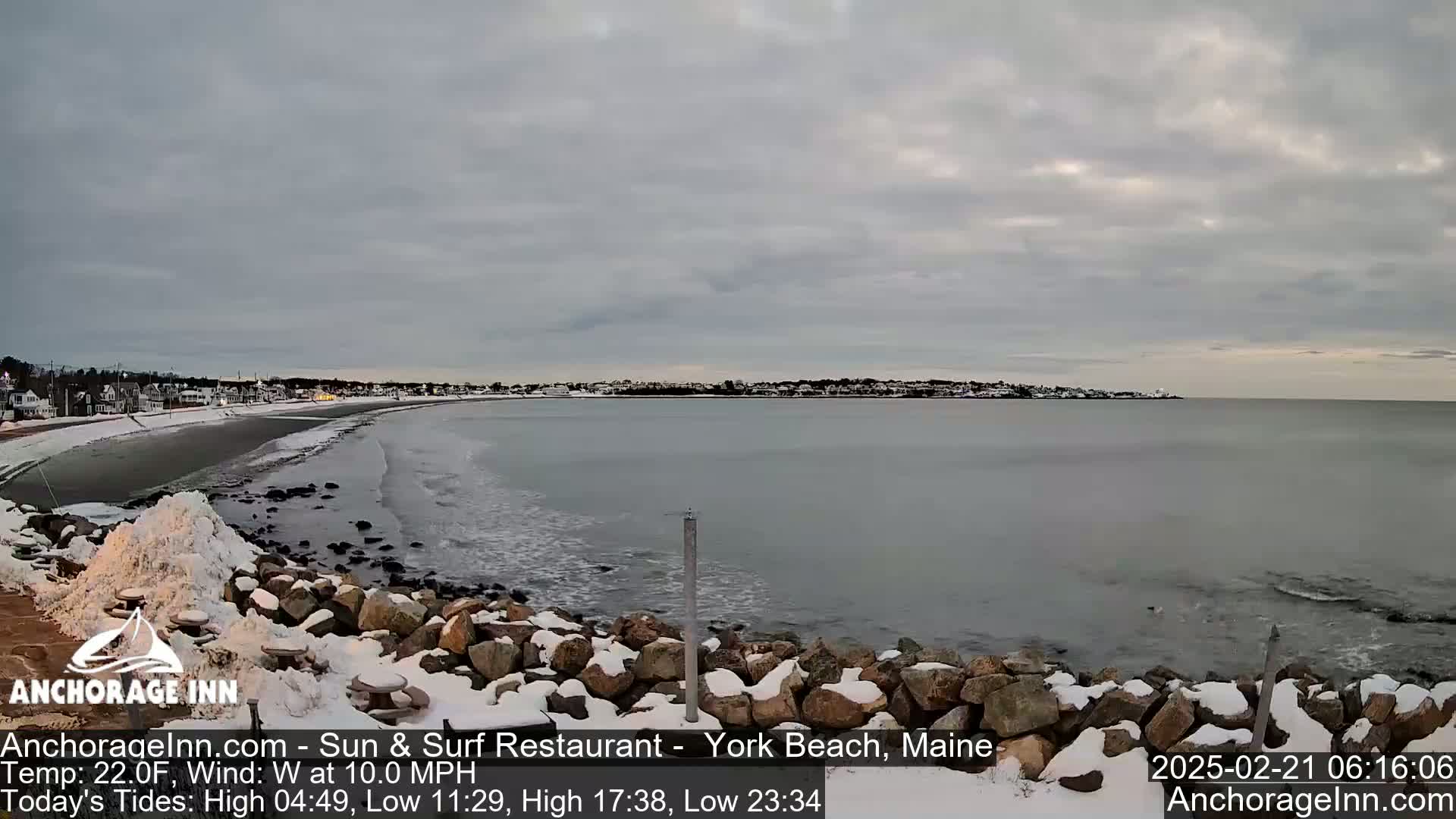 A snow-dusted rocky shoreline and beach overlook a calm, gray ocean under an overcast sky.