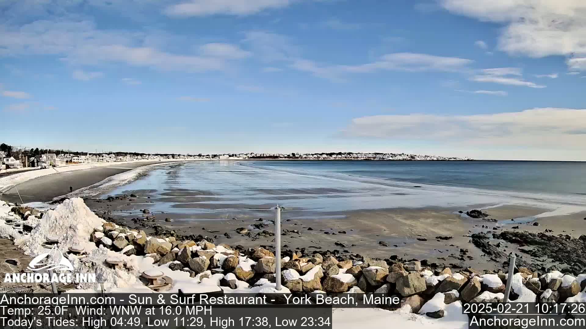 A partially frozen beach with snow-covered rocks in the foreground and a town visible in the distance under a mostly sunny sky.