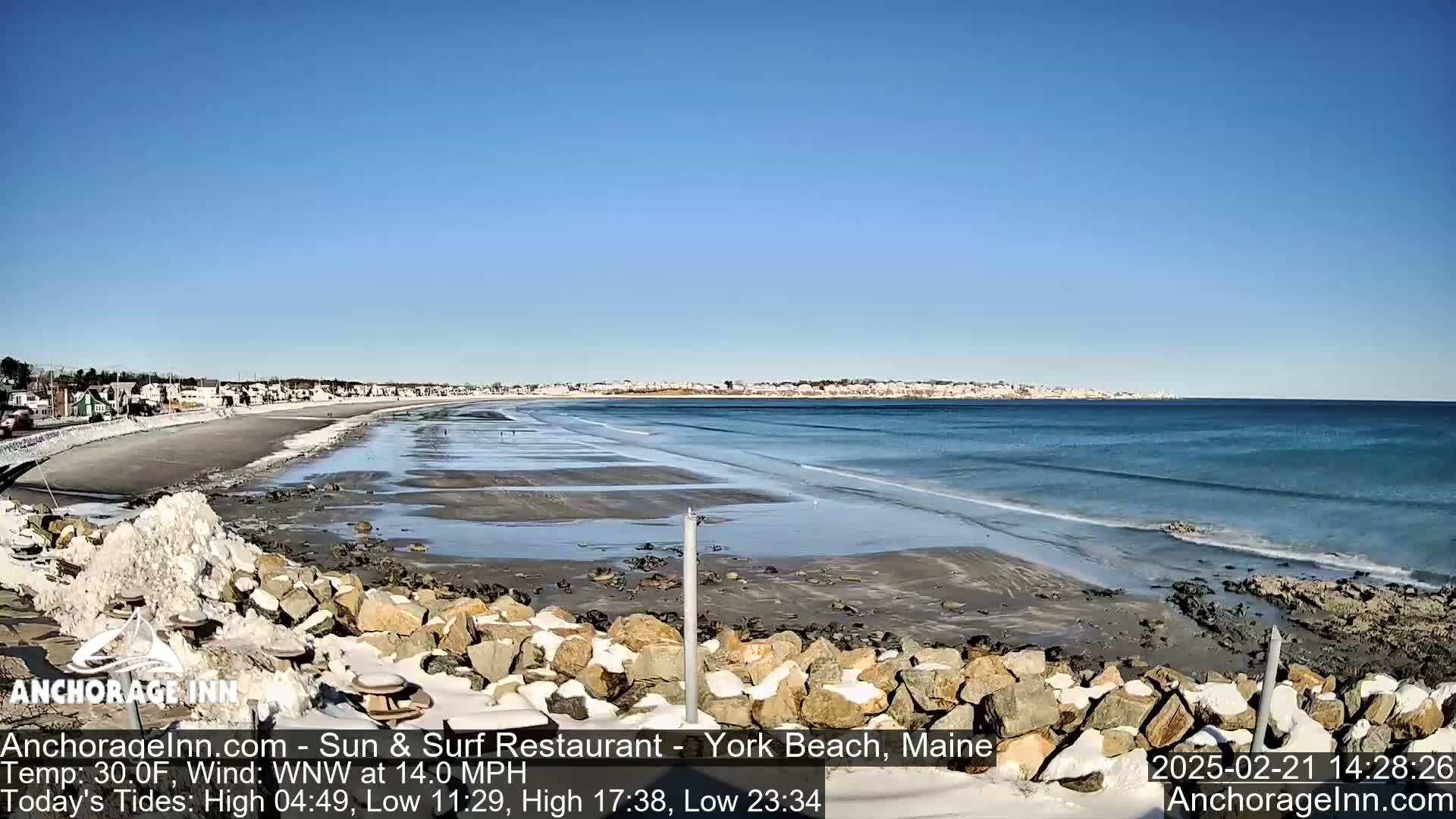 A mostly sunny day at a snowy beach shows a calm ocean with small waves lapping a wide sandy shore, backed by a town.