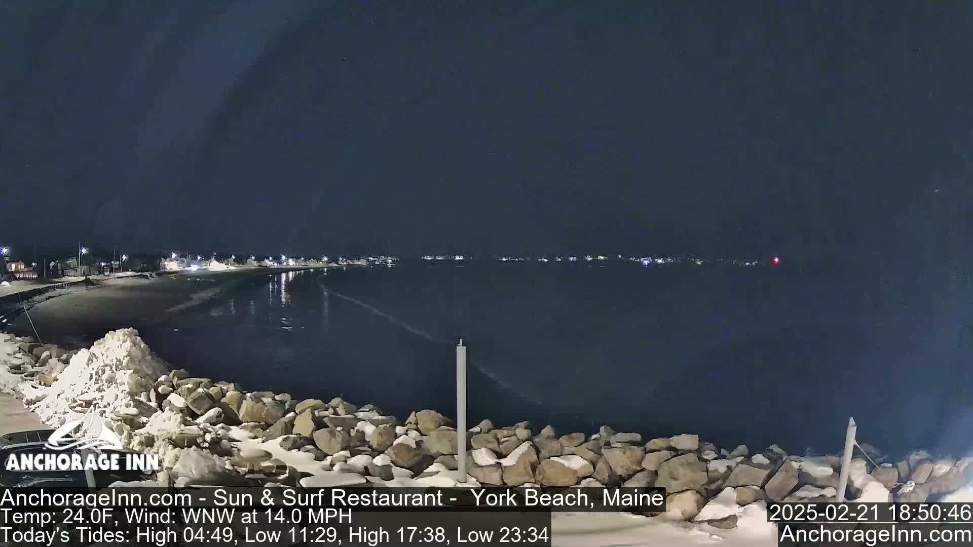 A nighttime view of a snow-dusted rocky shoreline and calm dark water, with a town faintly lit in the distance under a clear, dark sky.