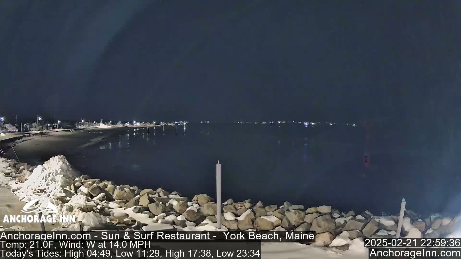 A nighttime view of a snow-dusted rocky shoreline and calm, dark water, with a town faintly lit in the distance under a clear, dark sky.