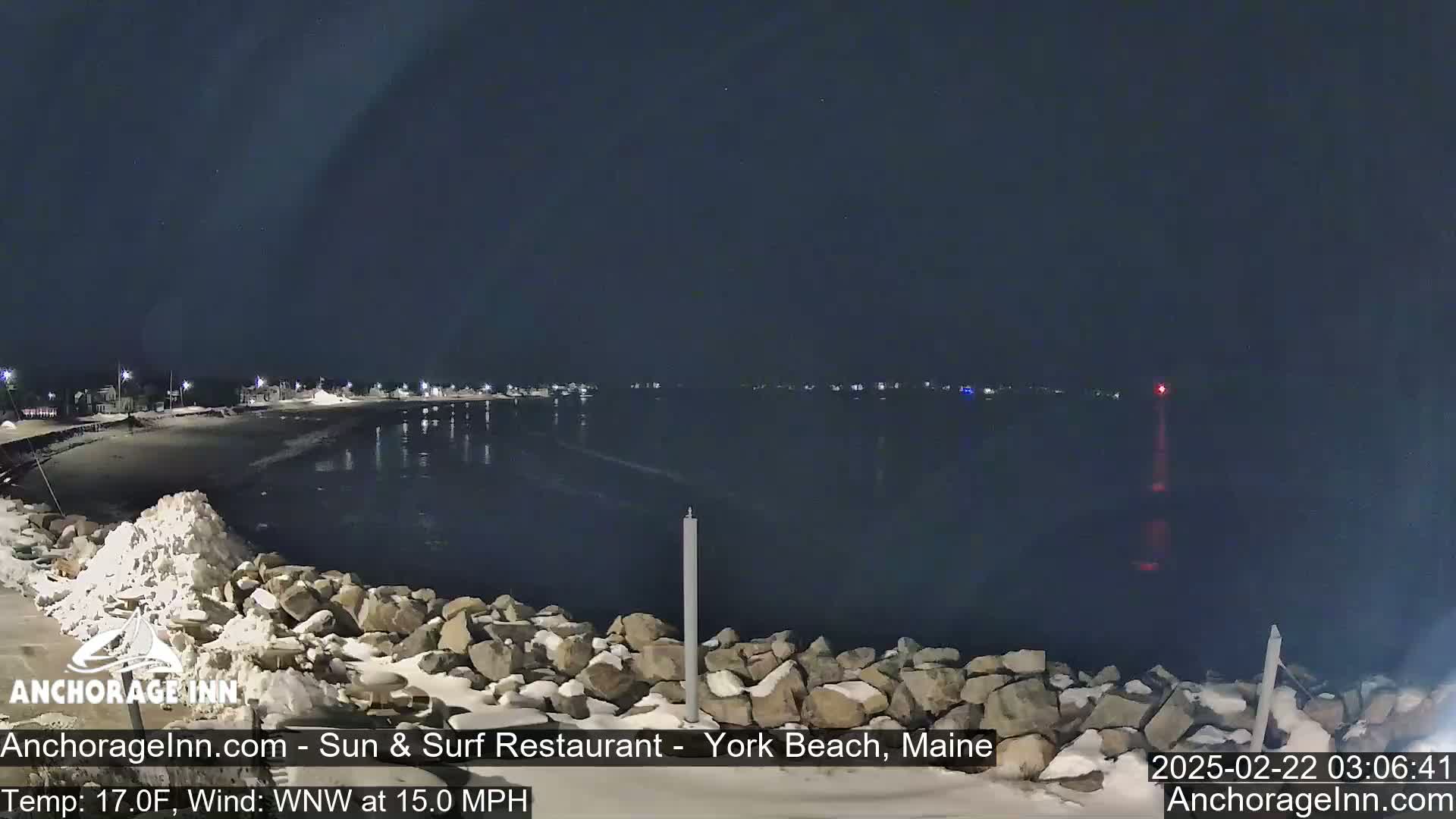 A nighttime view of a snow-covered rocky shoreline and calm dark water, with a town faintly lit in the distance under a clear, dark sky.