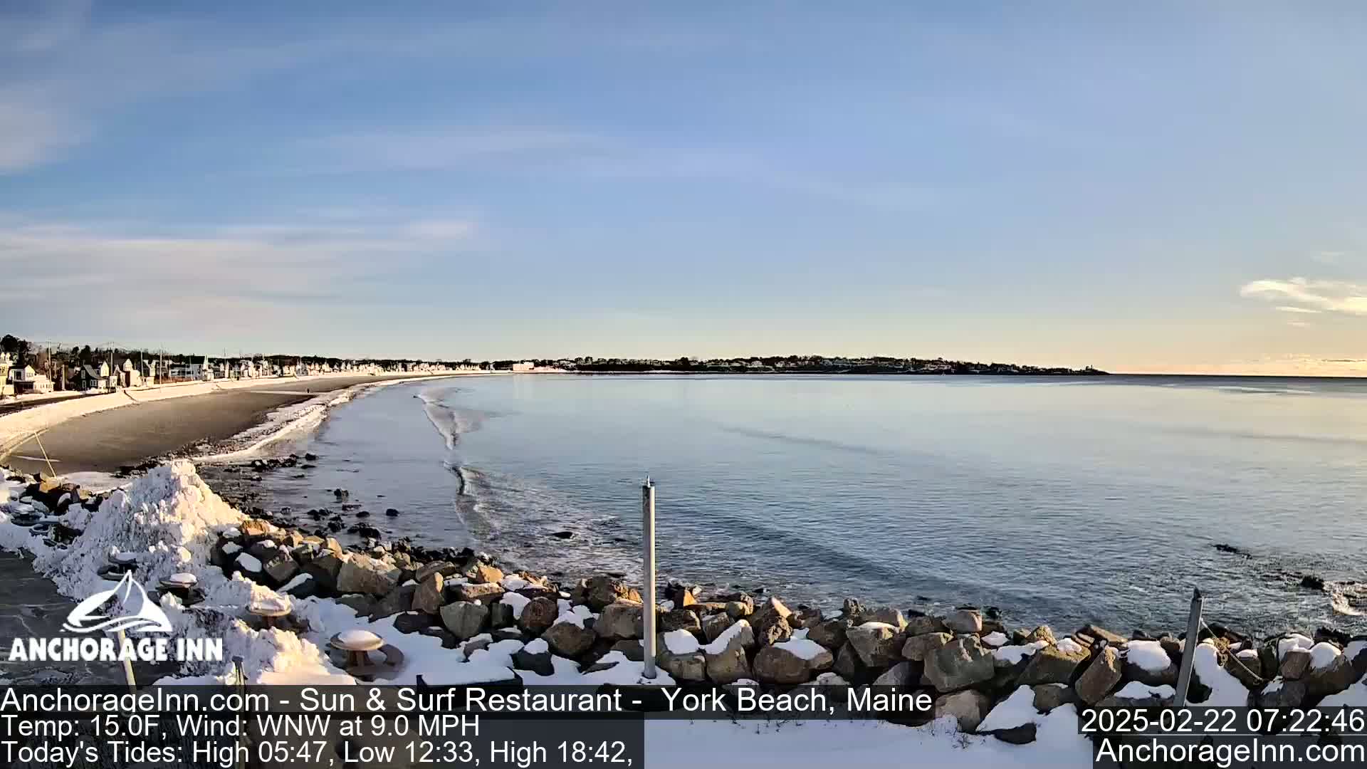 A snow-covered rocky shoreline overlooks a calm ocean with a sandy beach and distant houses under a clear, bright sky.