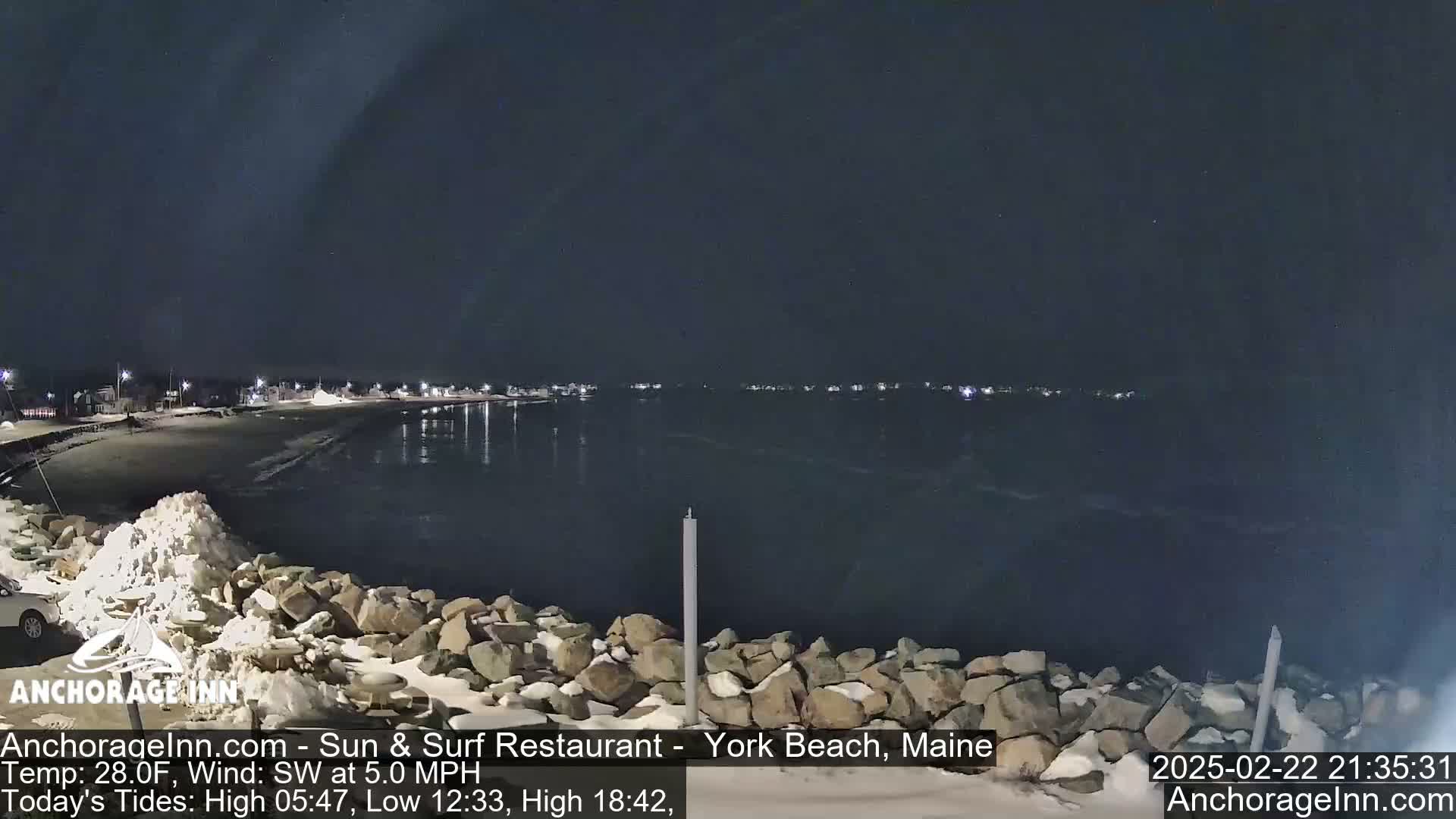 A nighttime view of a snow-covered rocky shoreline and a calm, dark body of water, with a town faintly visible in the distance under a clear sky.