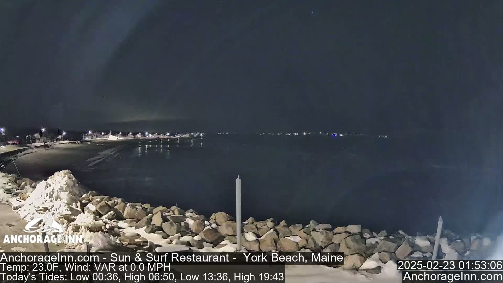 A nighttime view of a snow-dusted rocky shoreline and calm dark water, with a town faintly lit in the distance under a dark sky.