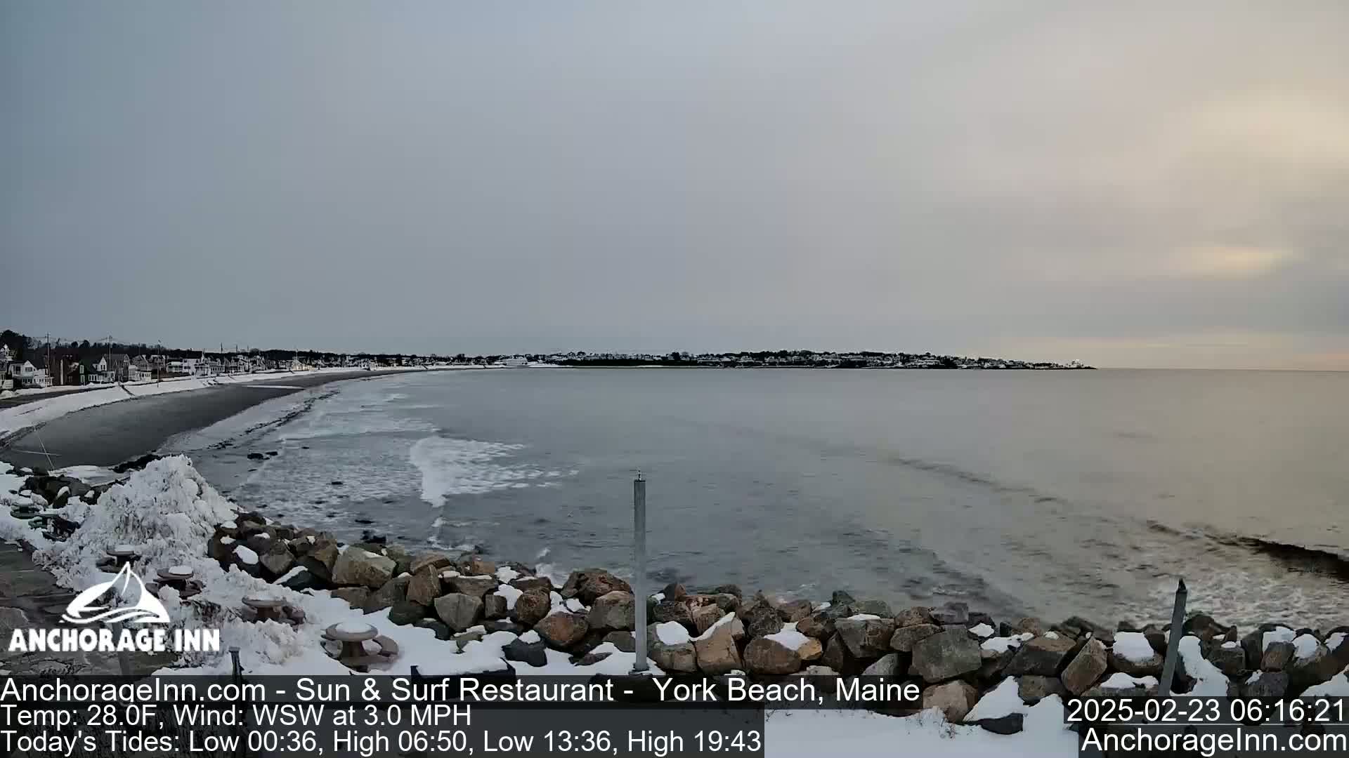 A snow-covered rocky shoreline meets a calm, gray ocean under an overcast sky.