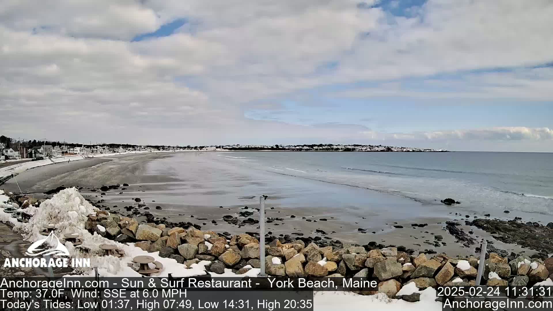 A partly cloudy day shows a snow-dusted beach with calm waves lapping the shore, backed by a line of houses.