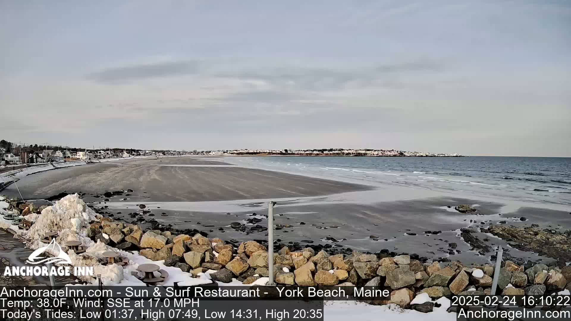 A partially frozen beach with a rocky shoreline and a town in the distance under a cloudy sky.