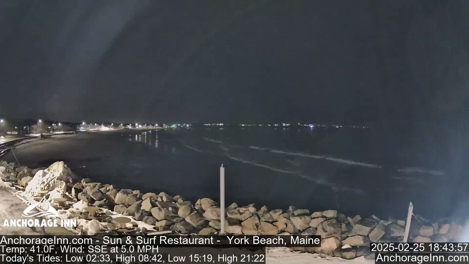 A nighttime view of a rocky shoreline and calm ocean with a town's lights visible in the distance under a dark sky.
