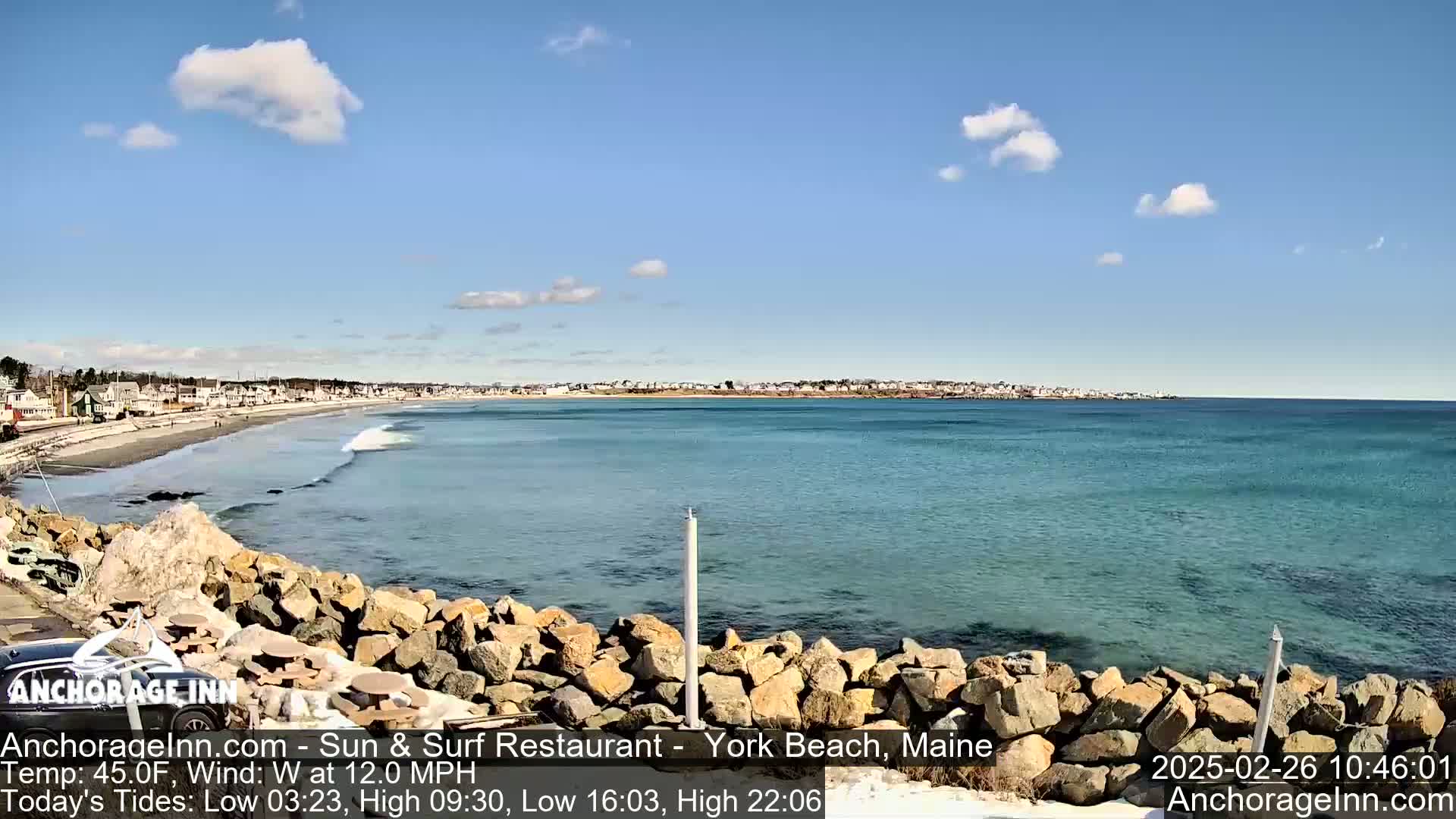 A rocky shoreline overlooks a calm, teal ocean with small waves lapping a sandy beach lined with houses under a mostly sunny sky.