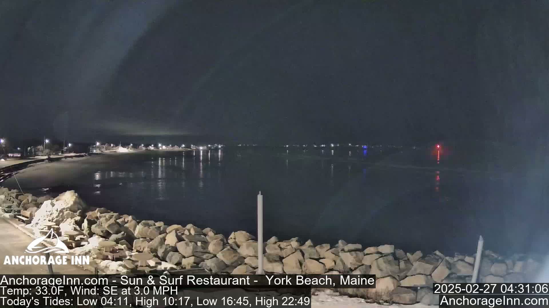 A nighttime view of a calm body of water reflecting the lights of a distant shoreline, with a rocky embankment in the foreground under a dark sky.