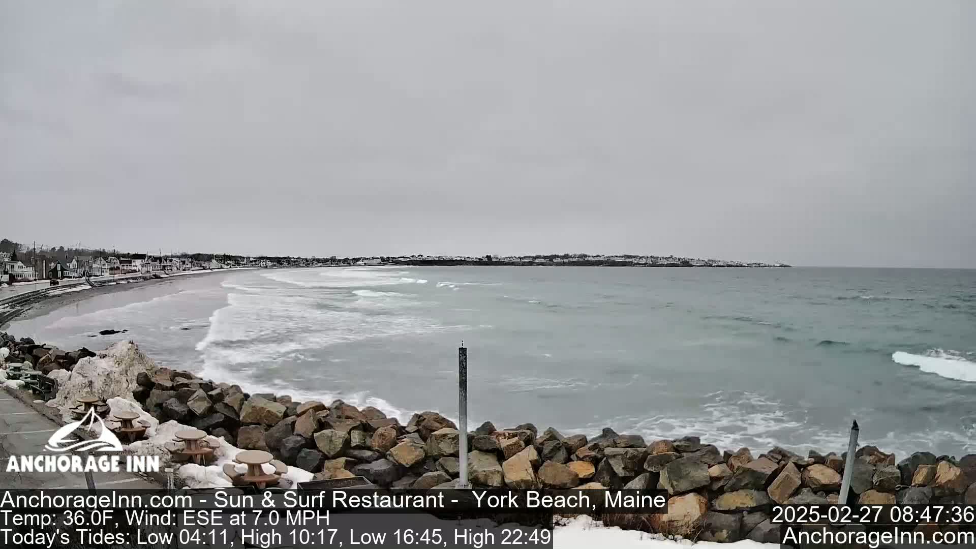 A rocky shoreline with a partially snowy beach and calm ocean waves stretches under an overcast sky.