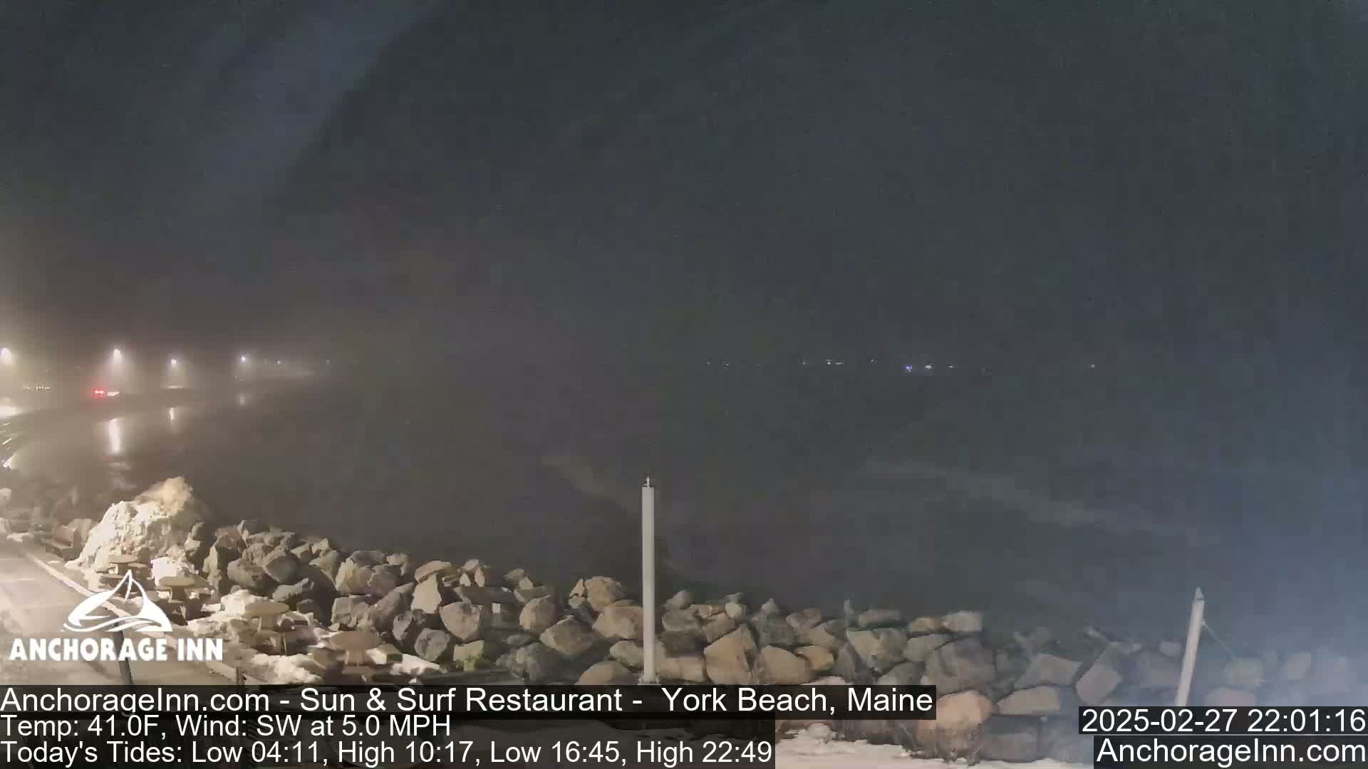 A foggy nighttime scene shows a rocky shoreline and a road with faint car lights in the distance.