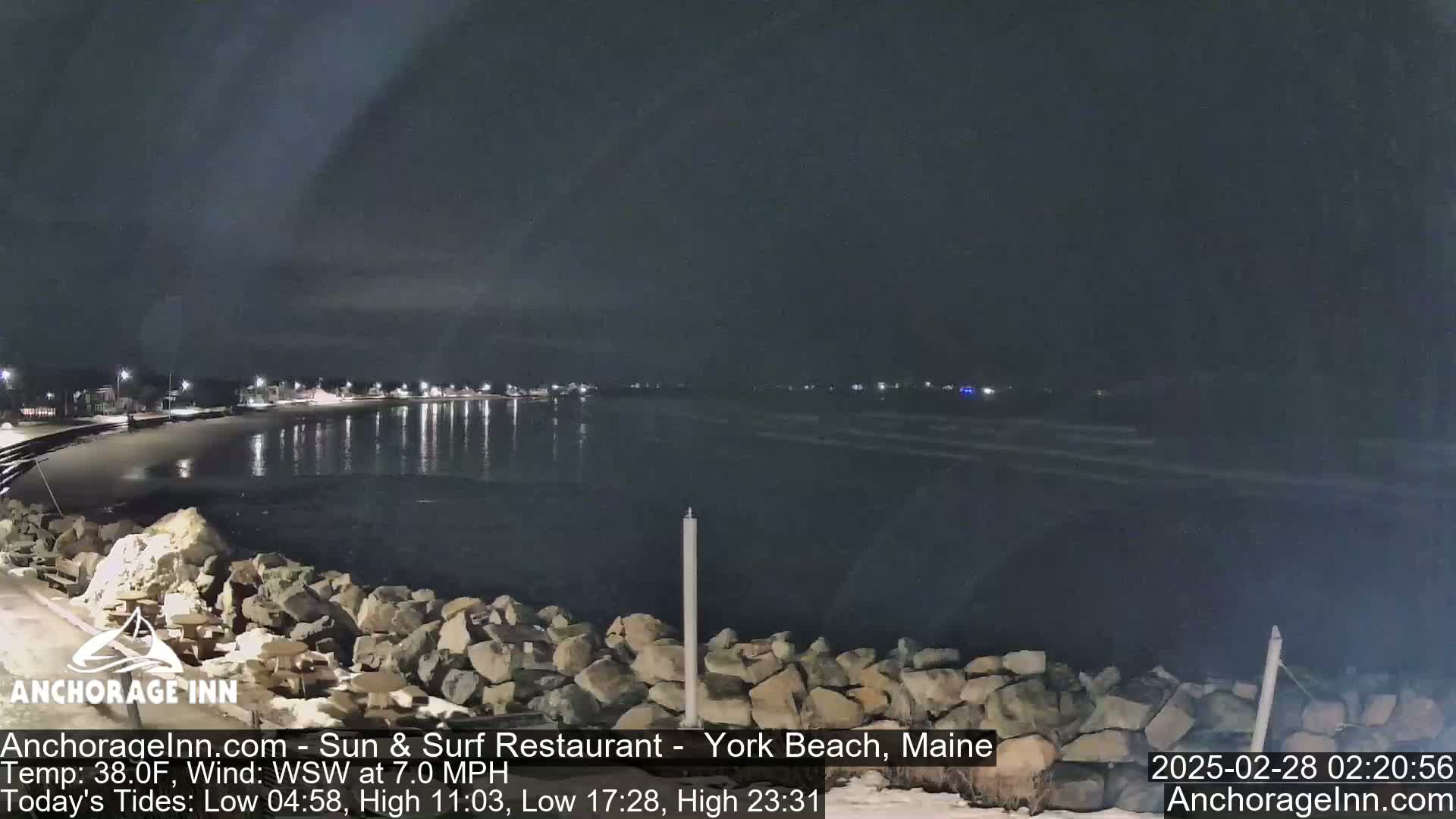 A nighttime view of a calm, dark ocean reflecting lights from a distant shoreline under a cloudy sky.