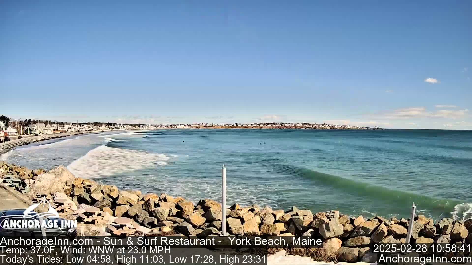 A rocky shoreline with gentle waves rolling onto a sandy beach under a clear blue sky.