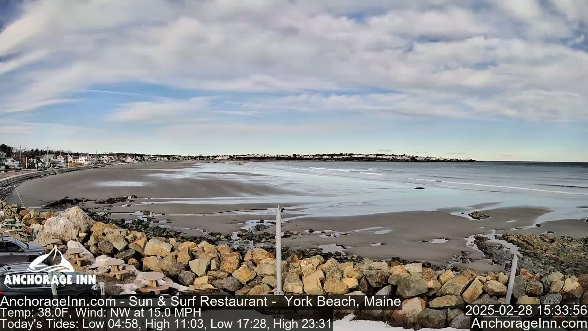 A sandy beach with a calm ocean under a partly cloudy sky is viewed from behind a low rock wall.