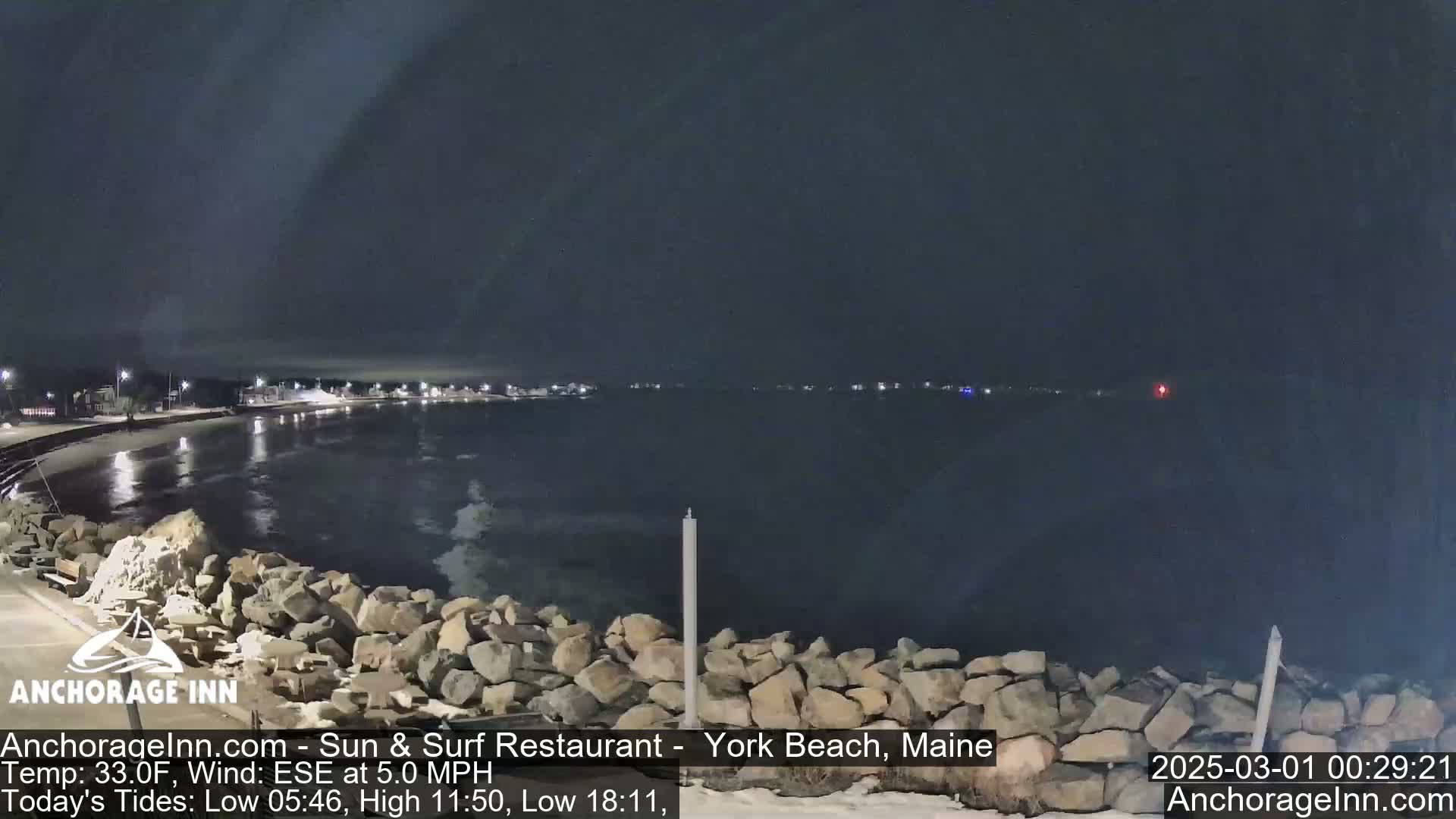 A nighttime view of a rocky shoreline and calm dark water, with distant lights visible across the bay under a cloudy sky.