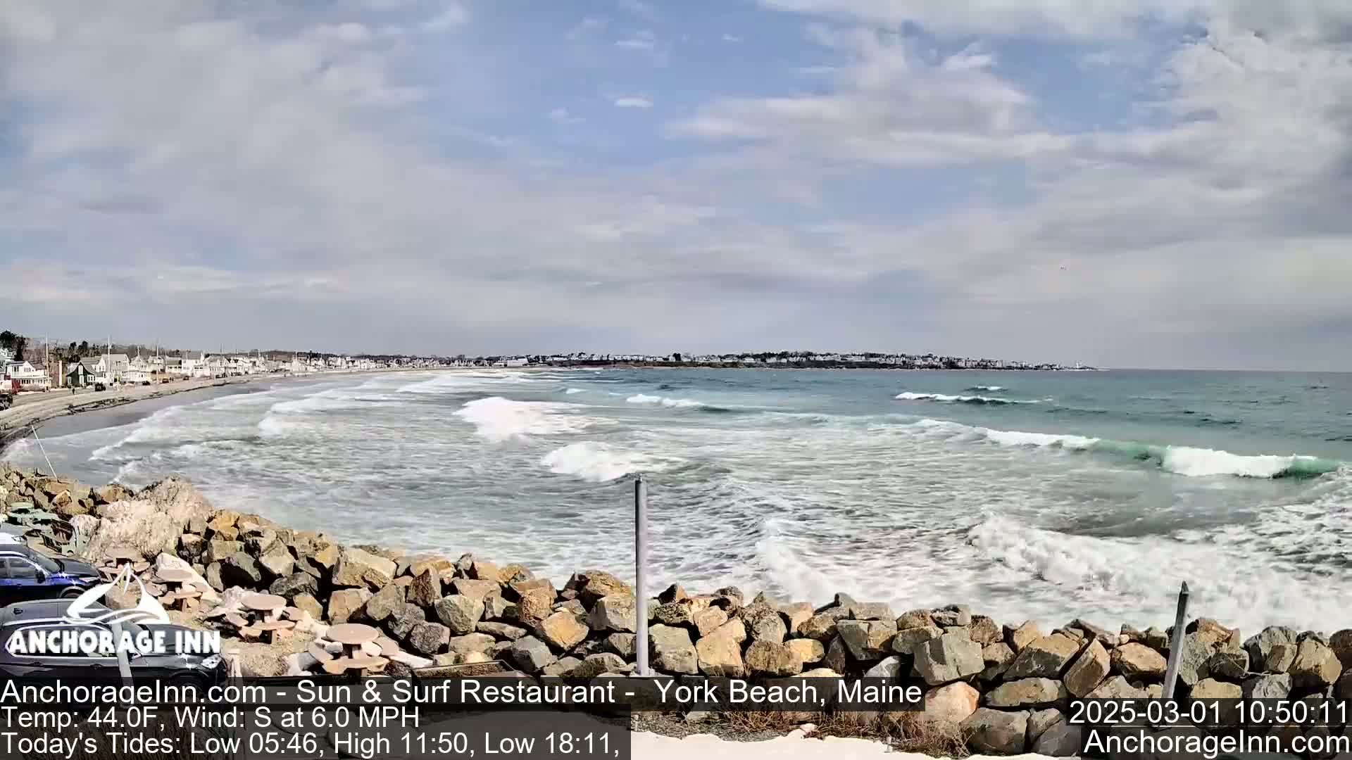 A partly cloudy day shows a rocky shoreline overlooking a beach with moderate waves crashing onto the sand.
