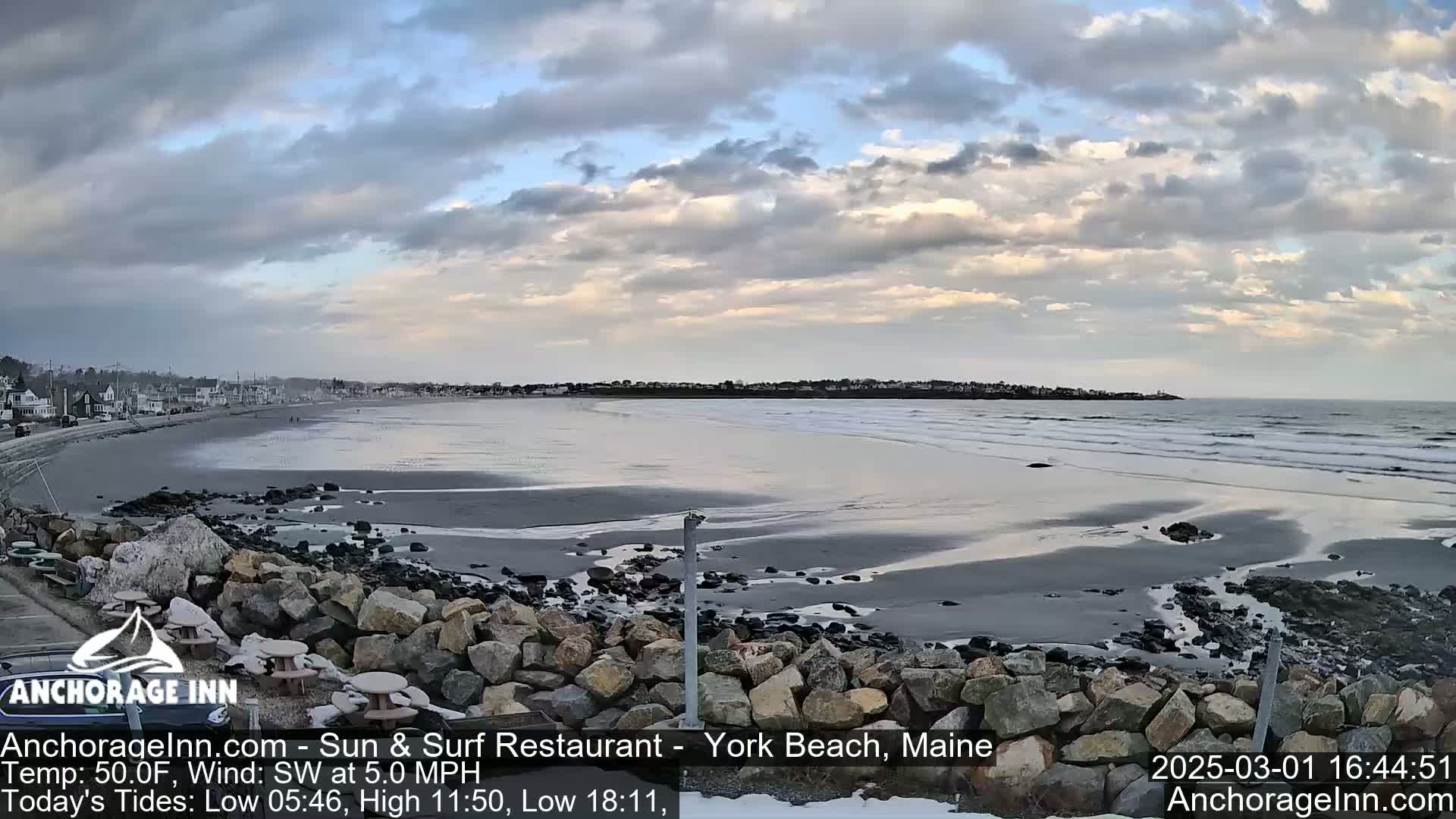 A wide sandy beach with small waves under a partly cloudy sky is viewed from behind a rocky wall.