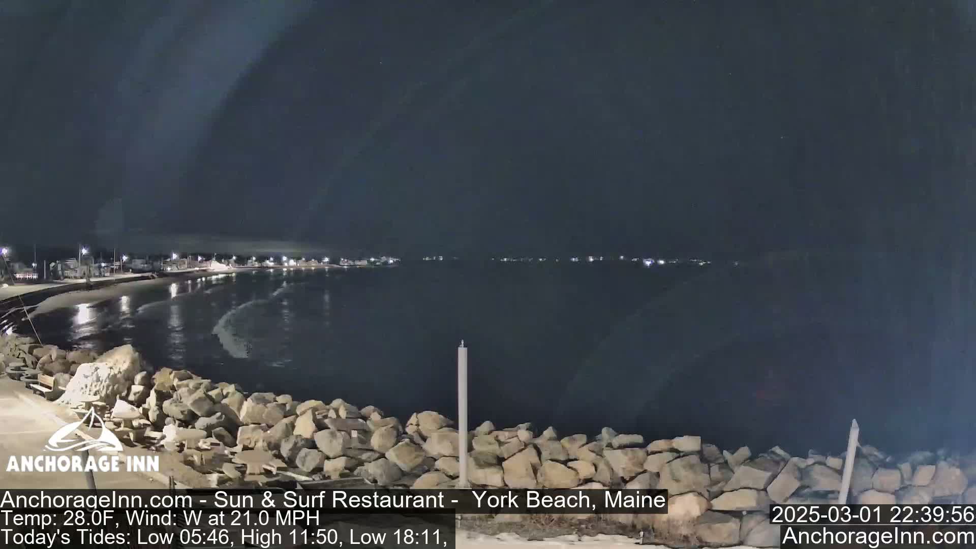 A nighttime view of a rocky shoreline and calm water, with faintly visible lights across the water under a dark sky.