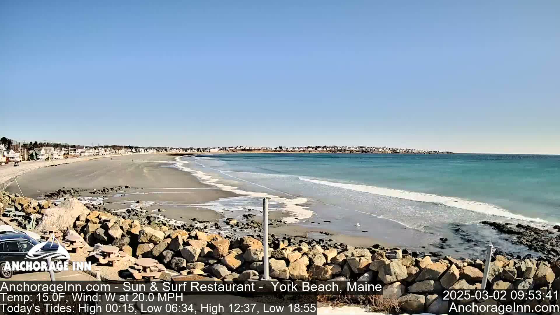 A sandy beach with a rocky shoreline stretches along a turquoise ocean under a clear blue sky, with a light wind.
