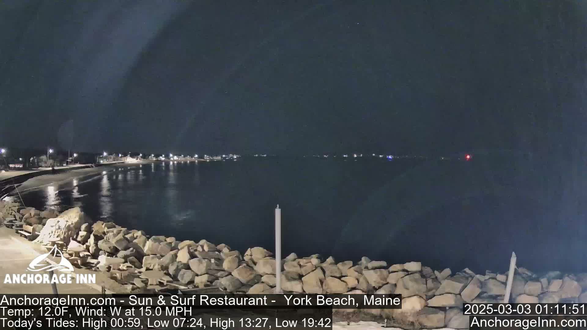 A nighttime view of a rocky shoreline and calm water, with a town faintly visible in the distance under a dark sky.