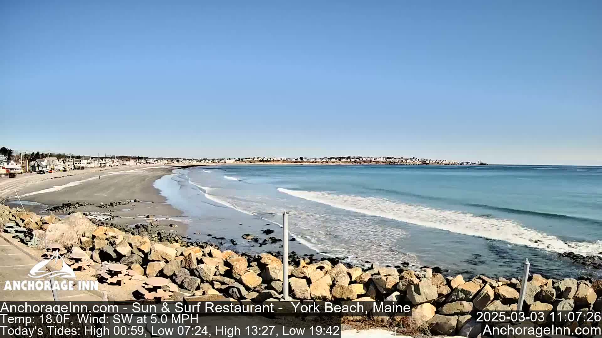 A long sandy beach with gentle waves under a clear blue sky is bordered by a rock wall in the foreground and houses in the distance.