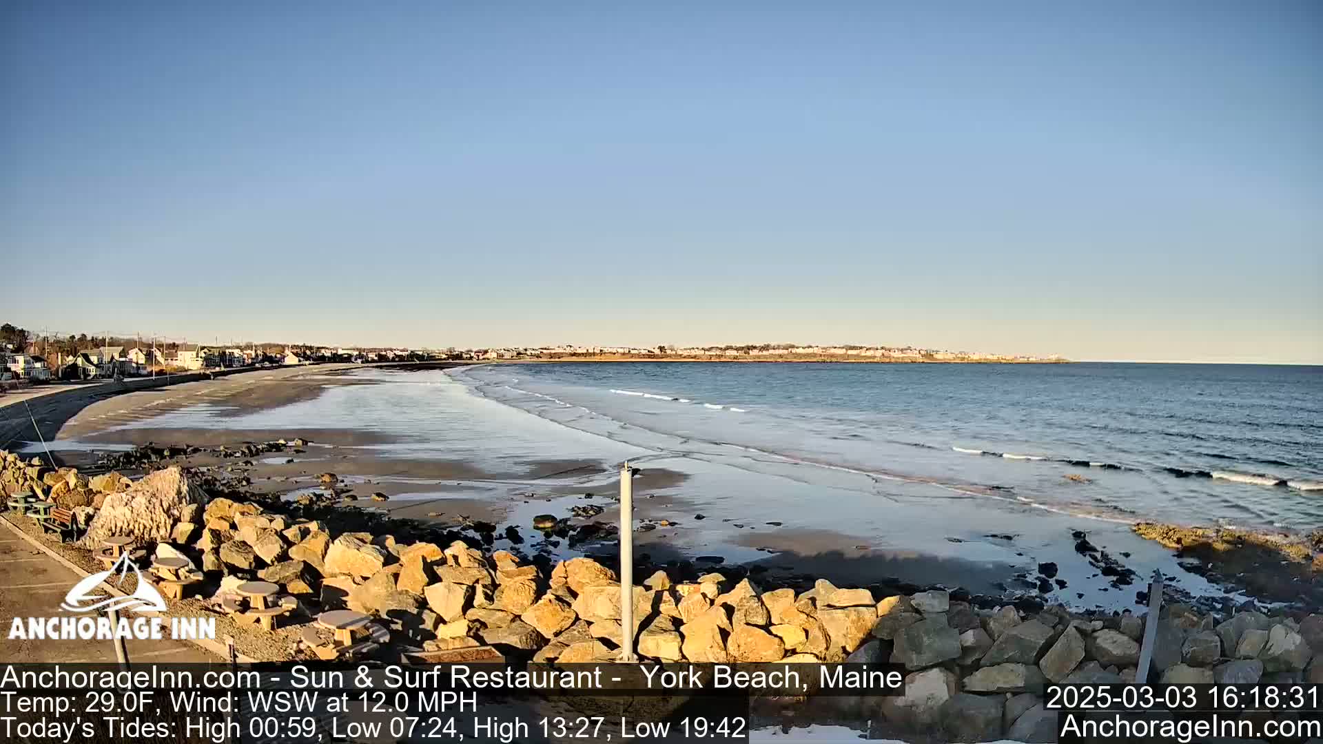 A sandy beach with gentle waves laps the shore under a clear blue sky, with a rocky embankment in the foreground and buildings visible in the distance.