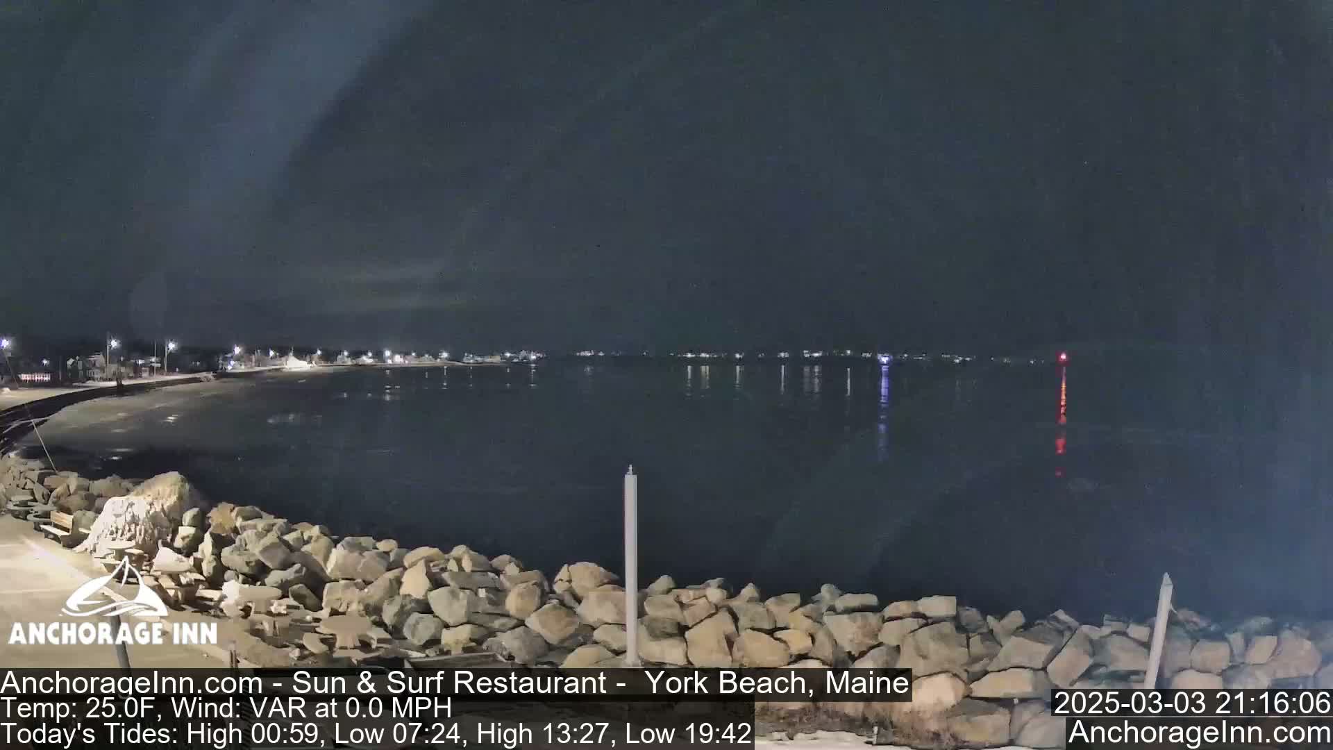 A nighttime, calm seascape shows a dark body of water reflecting the lights of a distant town under a clear, dark sky, with a rocky shoreline in the foreground.