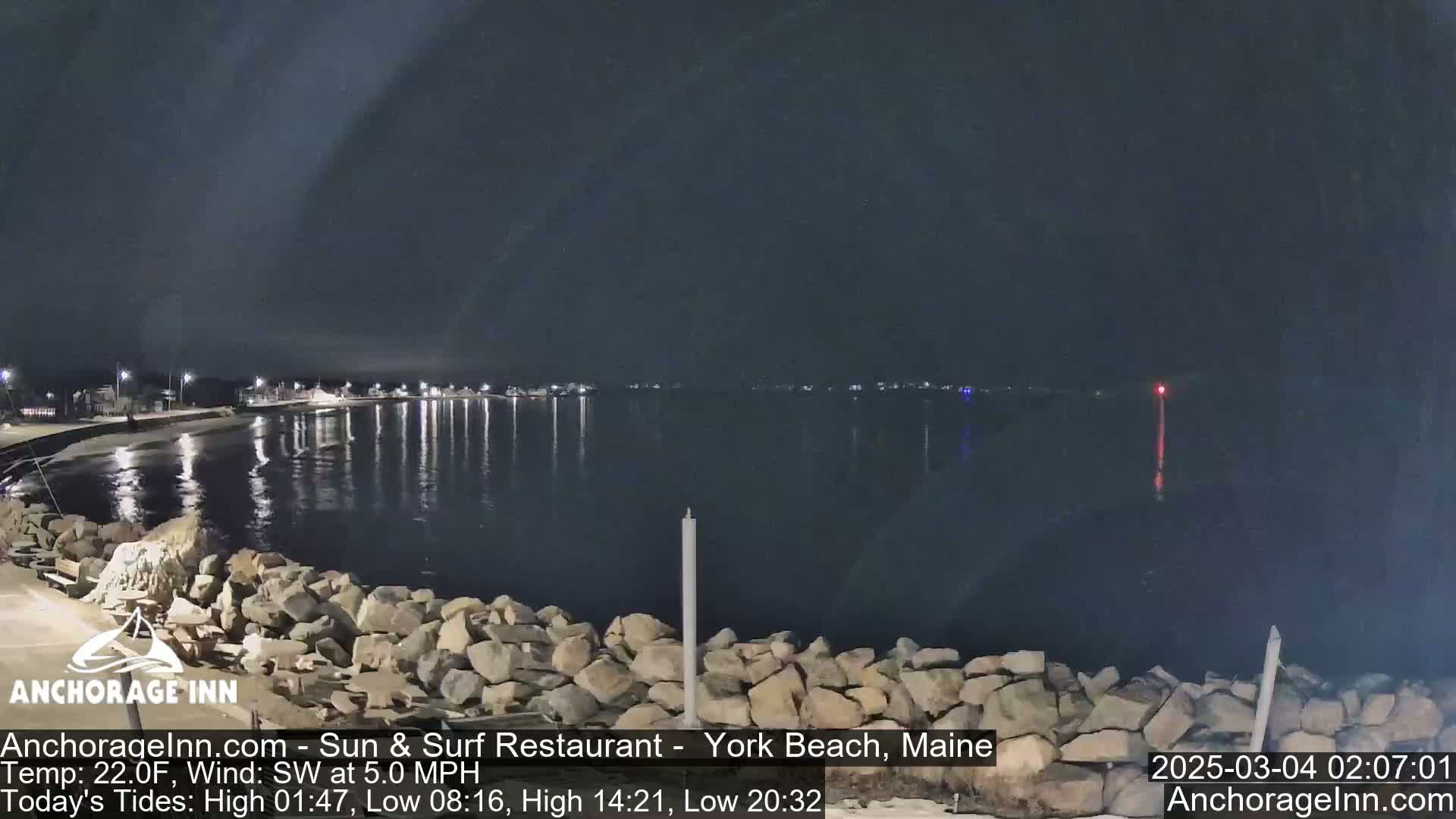 A nighttime view of a rocky shoreline and calm water, with lights visible across the water under a dark sky.
