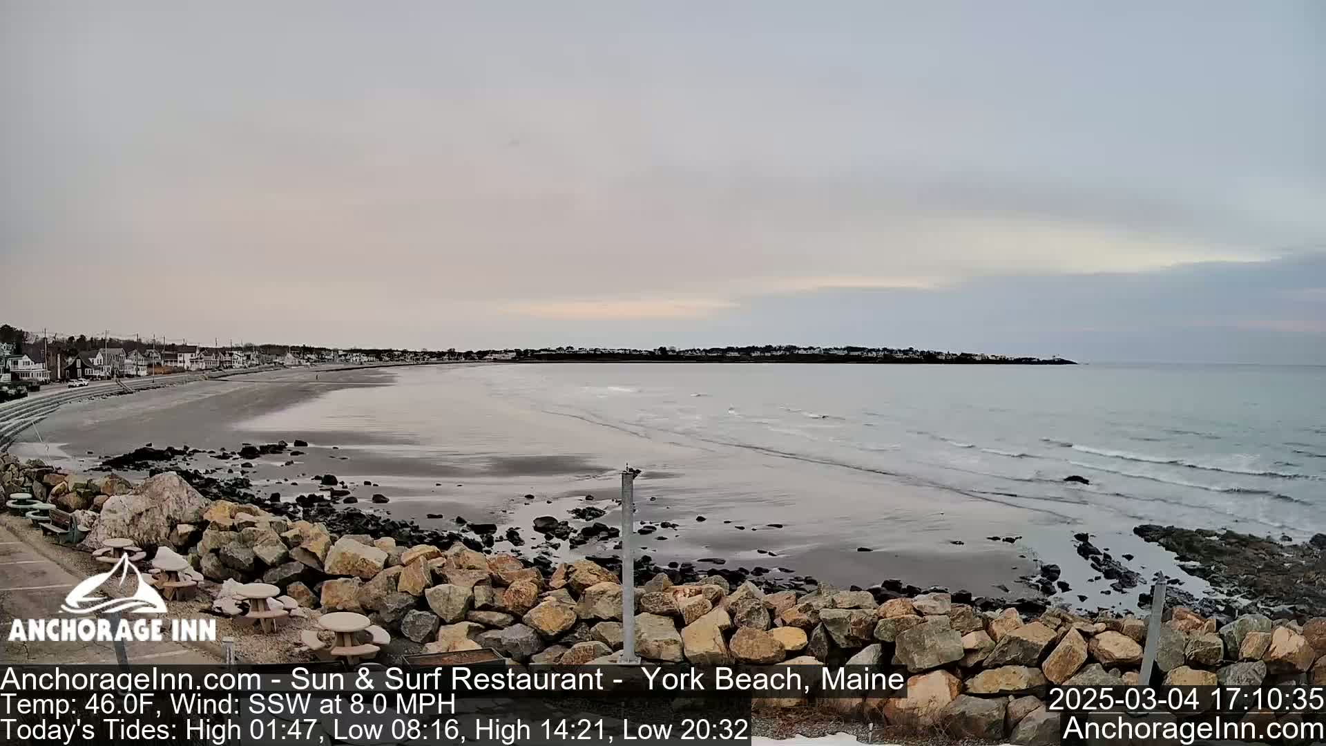 A mostly overcast sky hangs over a sandy beach with calm waves lapping the shore, backed by a row of houses.