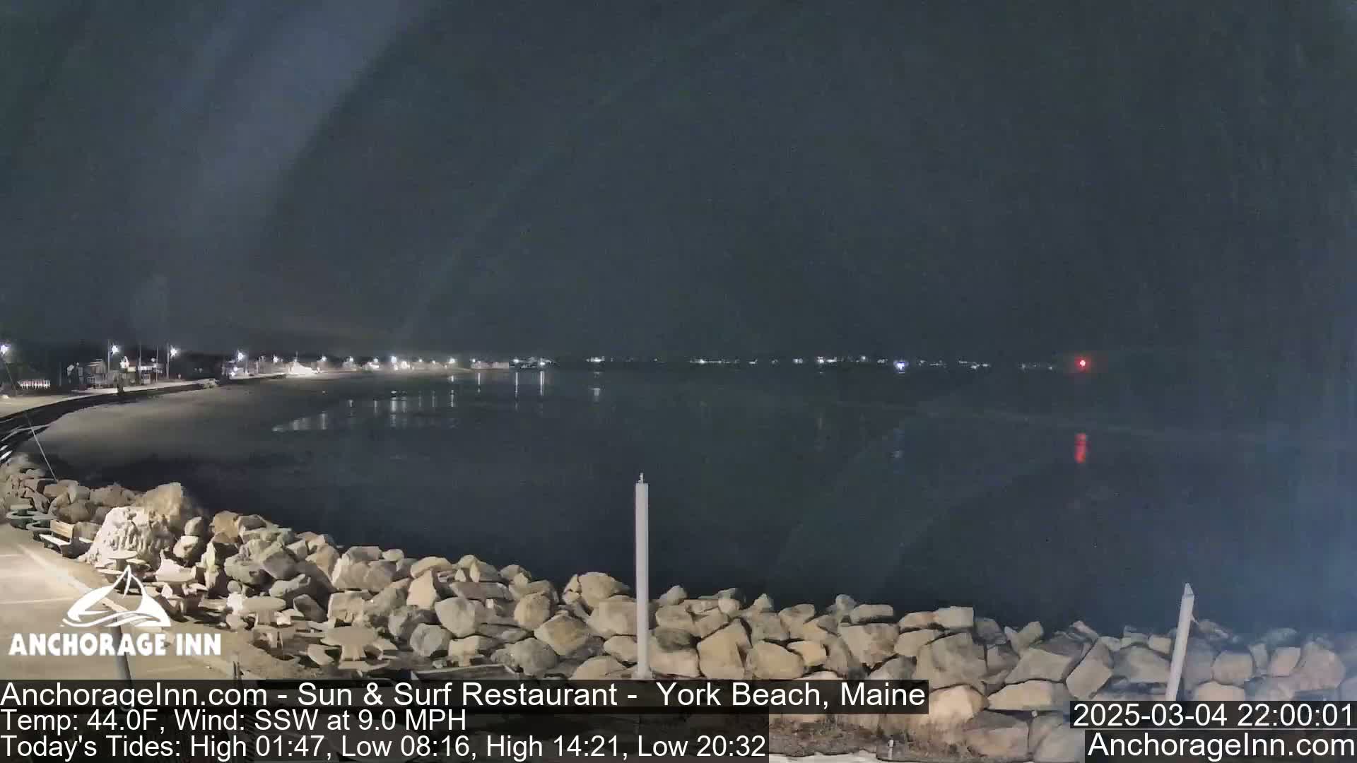A dark, nighttime view of a calm, reflective body of water next to a rocky shoreline and a dimly lit beachfront.