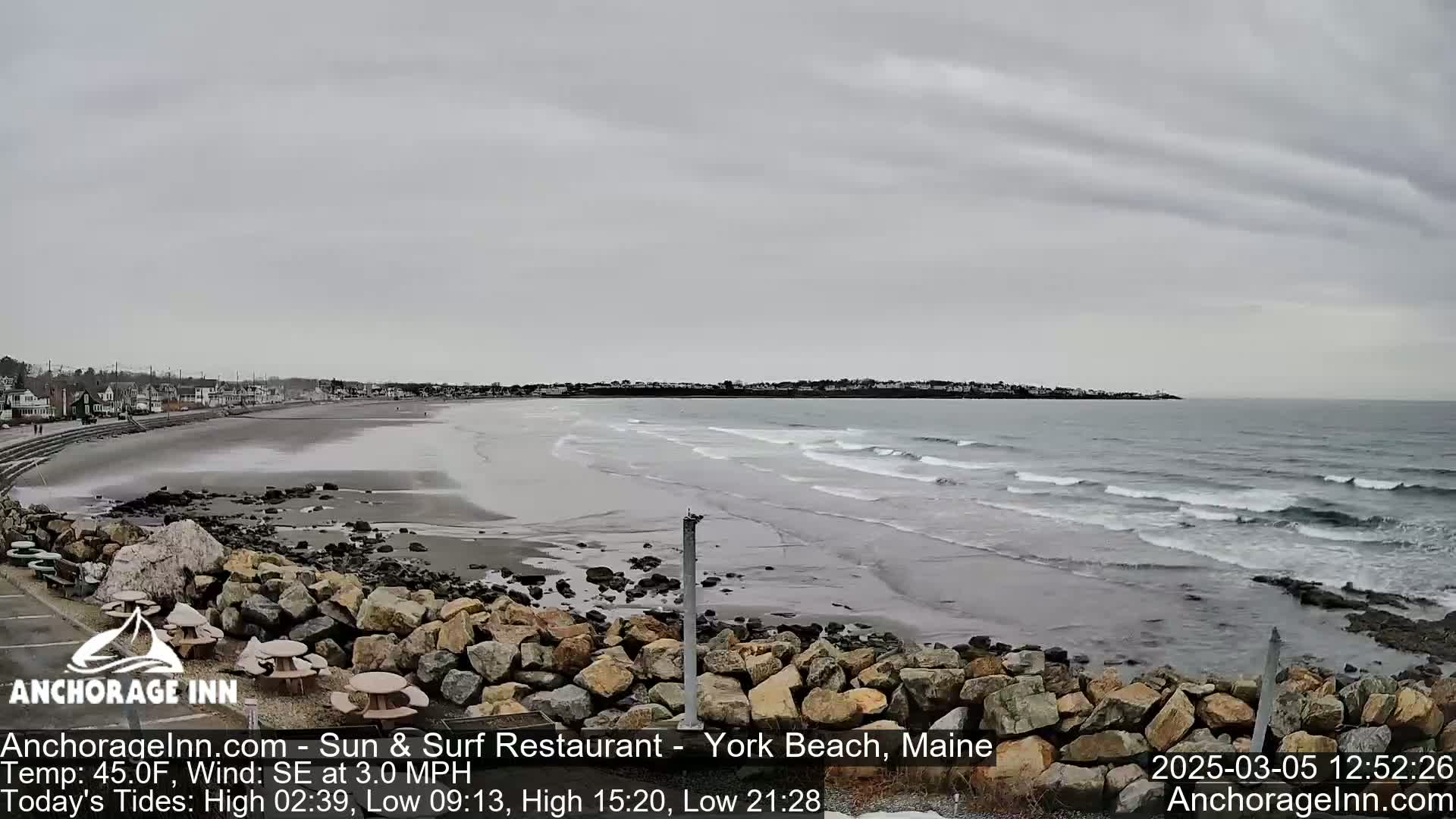 A gray, overcast day reveals a long sandy beach with gentle waves lapping the shore, backed by a rocky outcrop and a line of buildings in the distance.