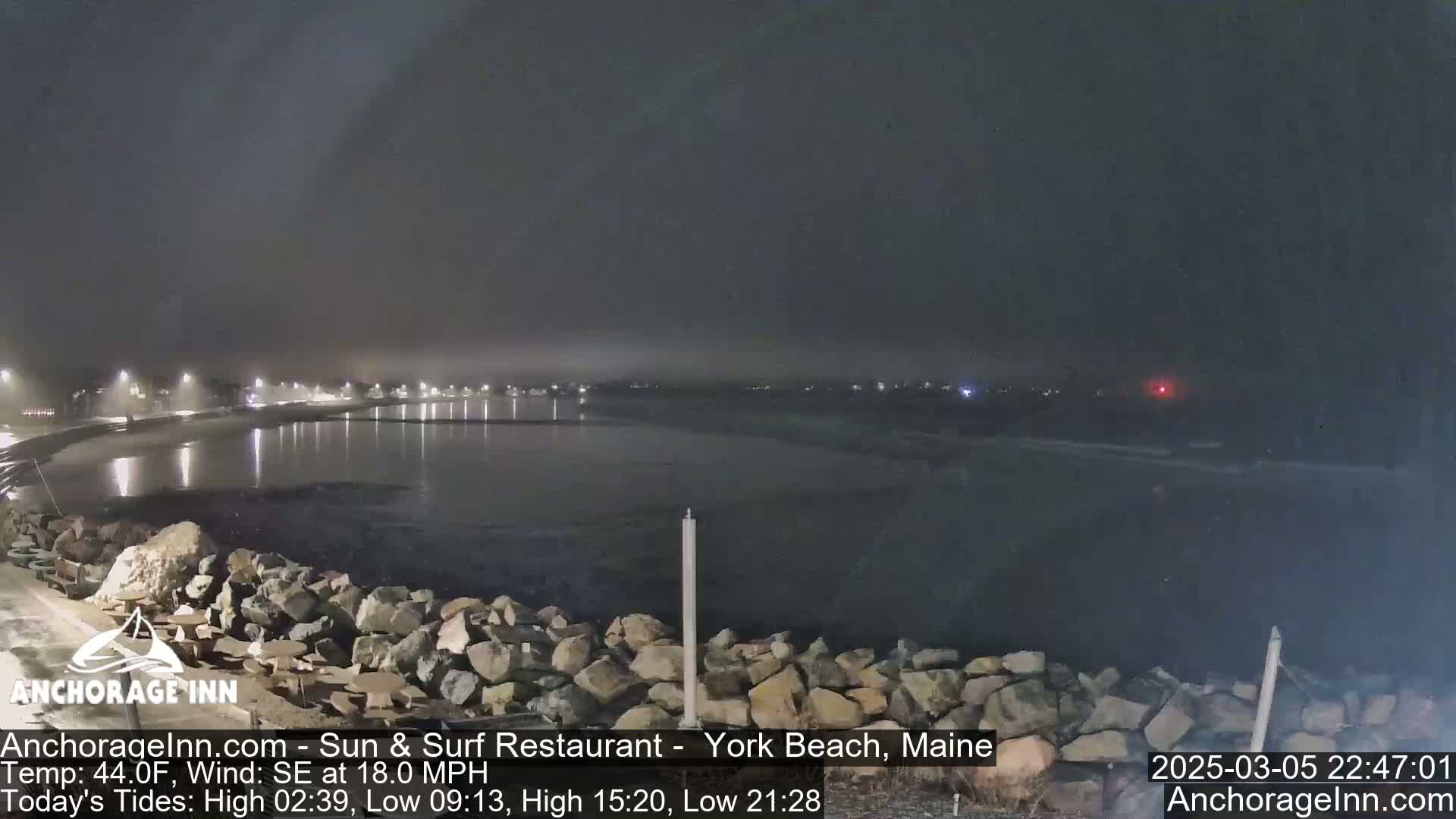 A nighttime coastal scene shows calm water reflecting lights from a distant shore under a hazy, dark sky.