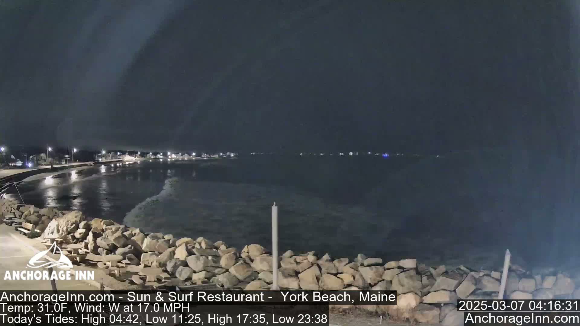 A nighttime view of a rocky shoreline and calm ocean under a dark, cloudy sky.