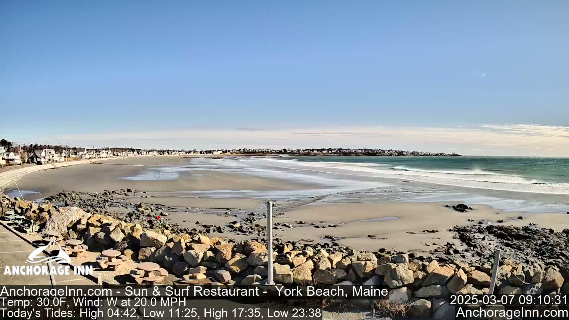 A sandy beach with small waves under a clear blue sky is seen from a rocky overlook, with houses visible in the distance.