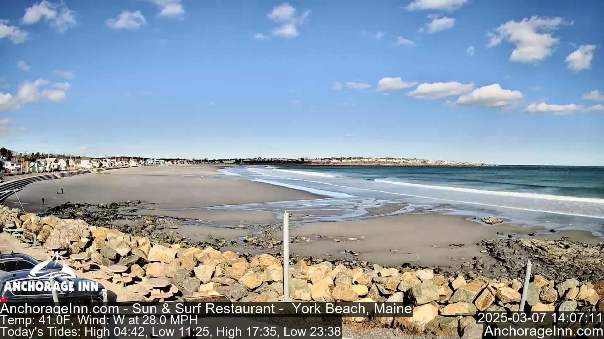 A sandy beach with waves rolling onto the shore under a partly cloudy sky is viewed from a rocky outcrop with a few buildings visible in the distance.