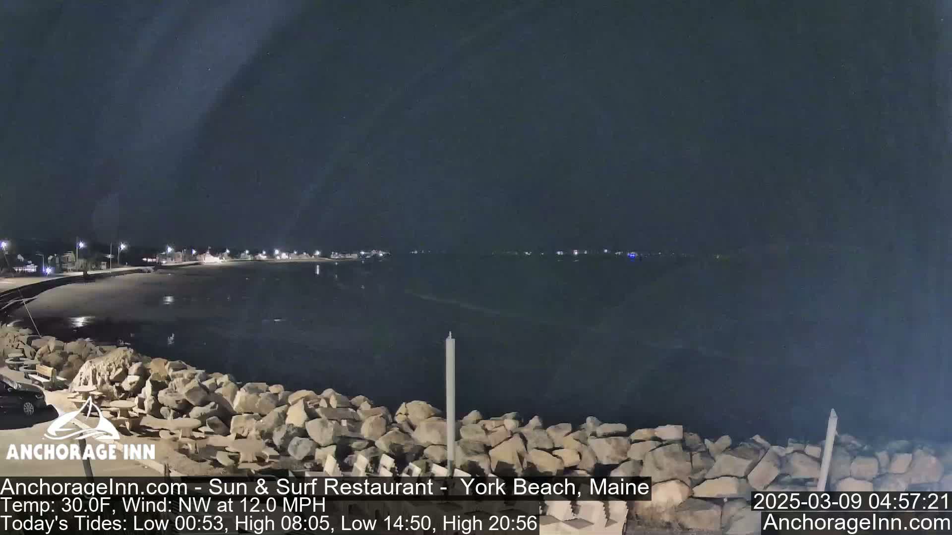 A nighttime view of a rocky shoreline and calm dark water, with a few lights visible in the distance under a clear sky.