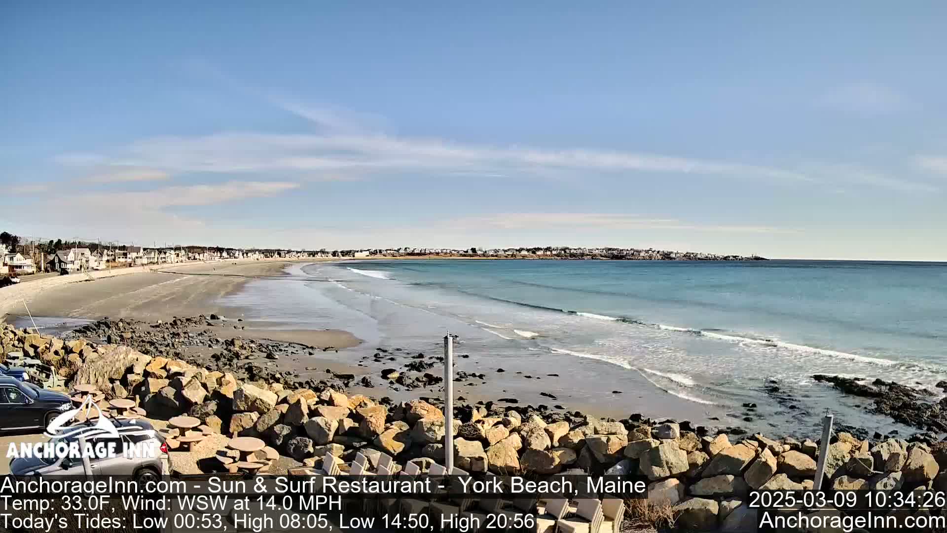 A sandy beach with gentle waves and a town in the background under a mostly clear sky.