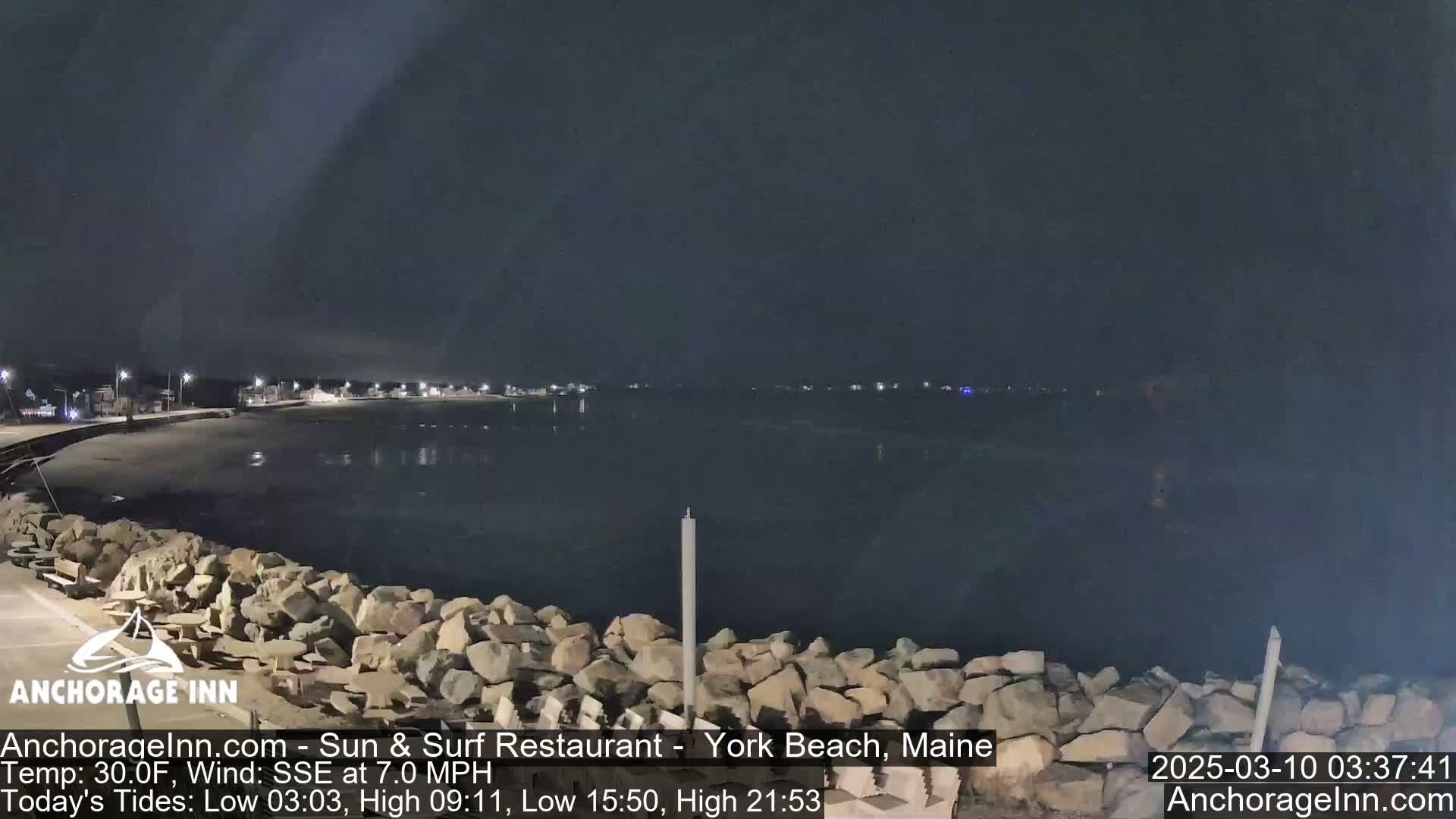 A nighttime view of a dark, calm ocean under a mostly clear sky, with a rocky shoreline and dimly lit buildings in the distance.