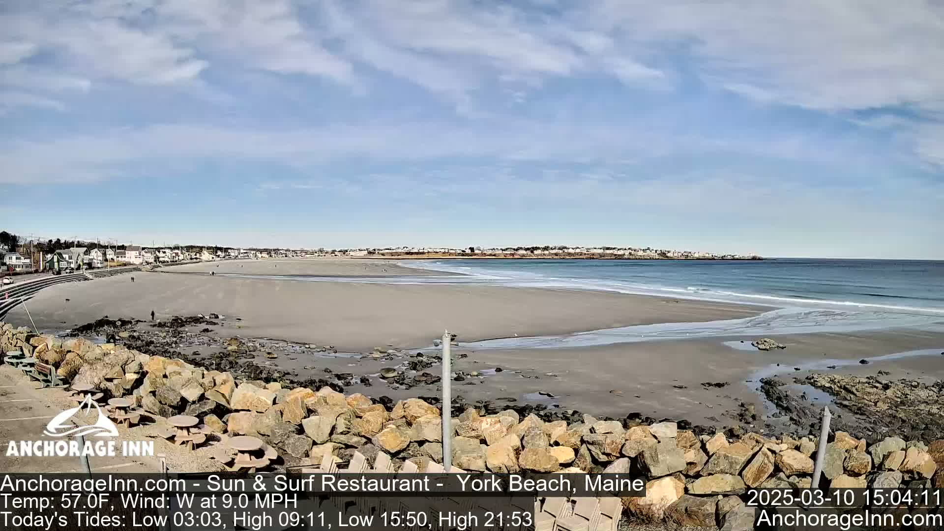 A sandy beach with gentle waves stretches out to a calm ocean under a mostly sunny sky.