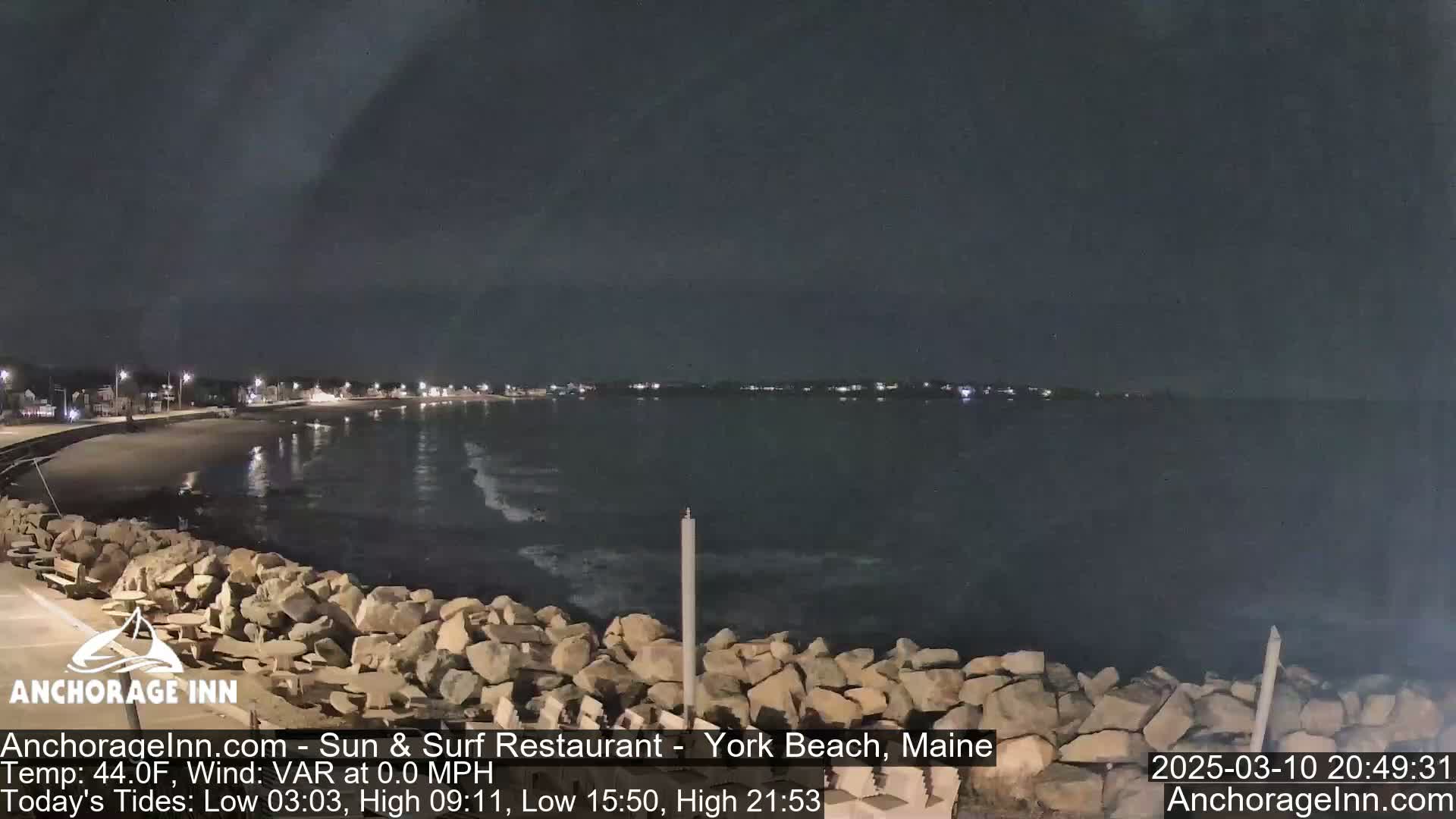 A nighttime view of a rocky shoreline and calm ocean, with a town faintly visible in the distance under a dark sky.