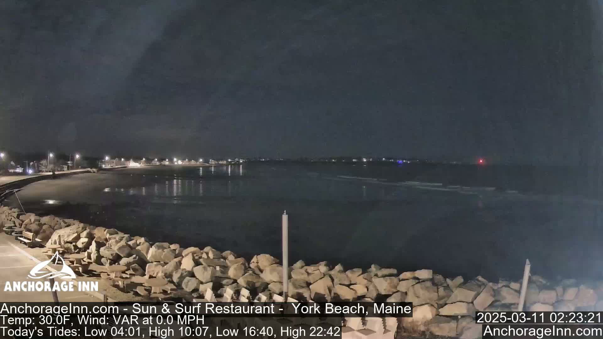 A nighttime view of a dark, calm ocean with a rocky shoreline and dimly lit buildings in the distance under a cloudy sky.