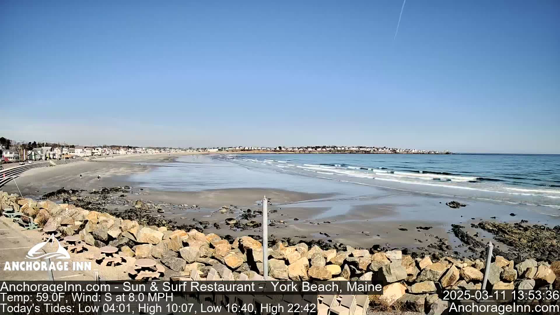 A sandy beach with small waves under a clear blue sky, viewed from behind a rocky embankment with outdoor seating.